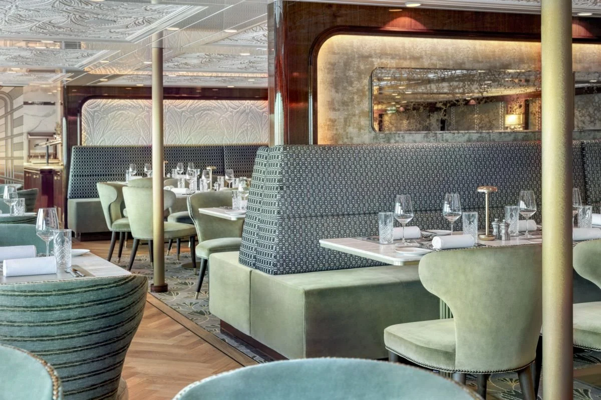 Elegant restaurant interior with green upholstered chairs, booth seating, and tables set with wine glasses, water glasses, napkins, and silverware.