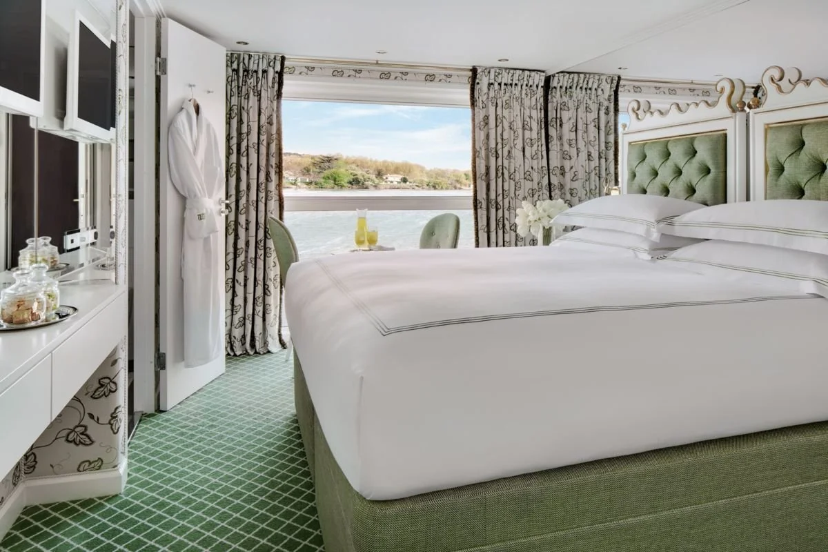 A hotel room with a large bed, green tufted headboard, and white linens, overlooking a river through a large window, with curtains and a small table with drinks.