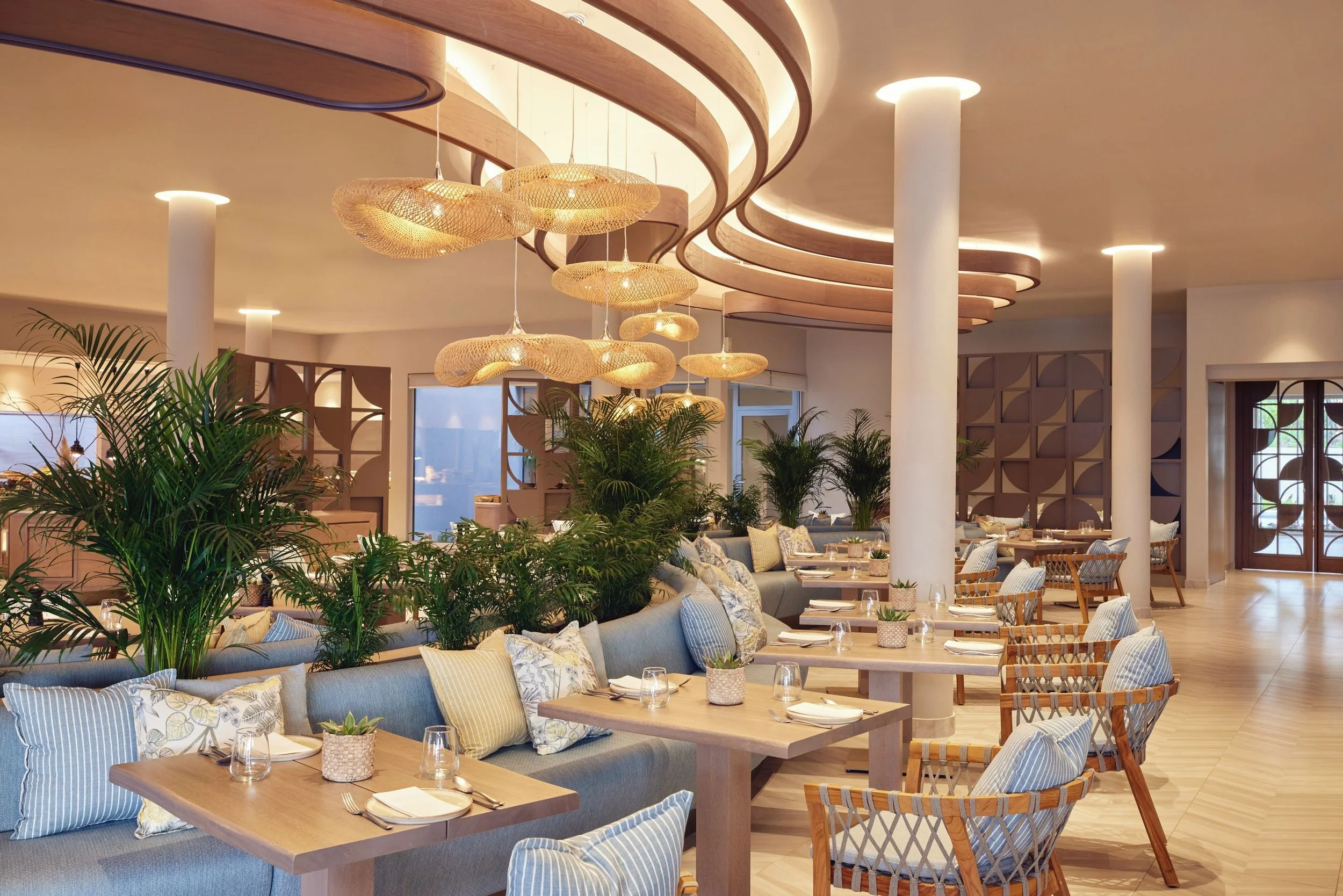 Restaurant interior decorated with string lights, plants, beige cushions, wooden tables, and a wooden ceiling with circular light fixtures.
