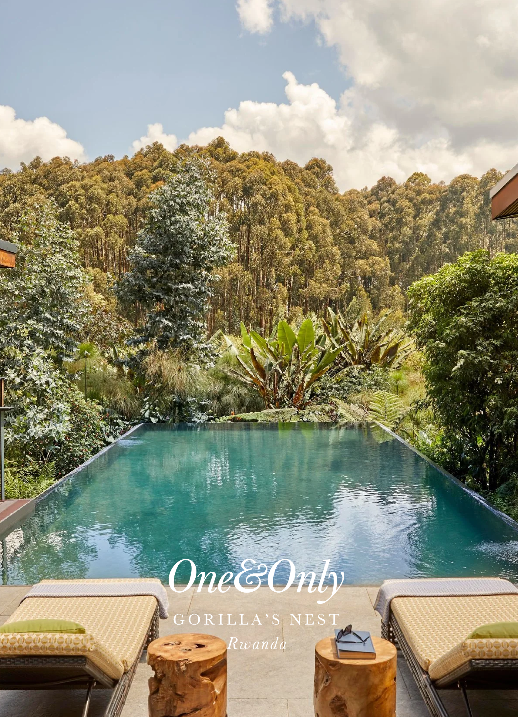 View of a swimming pool with outdoor lounge chairs and wooden stools, overlooking a lush green forest and a partly cloudy sky, labeled 'One&Only Gorilla's Nest Rwanda'.