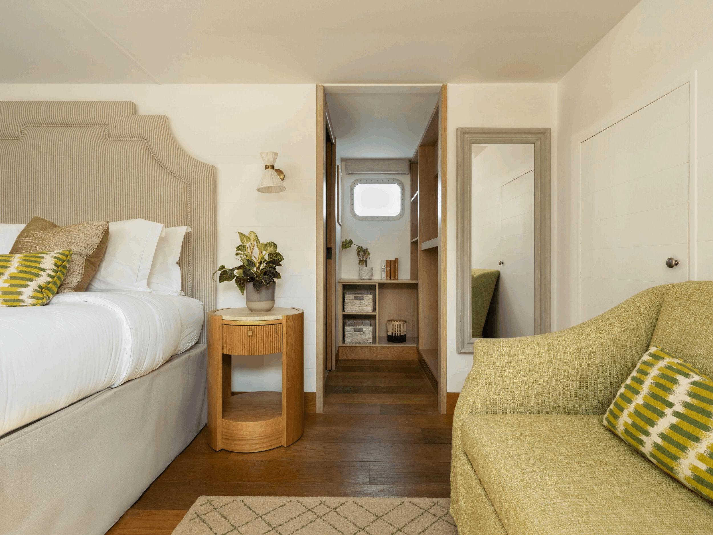 A cozy bedroom with a large bed featuring striped headboard and decorative pillows, a wooden side table with a plant, and a wooden bookshelf in the background.