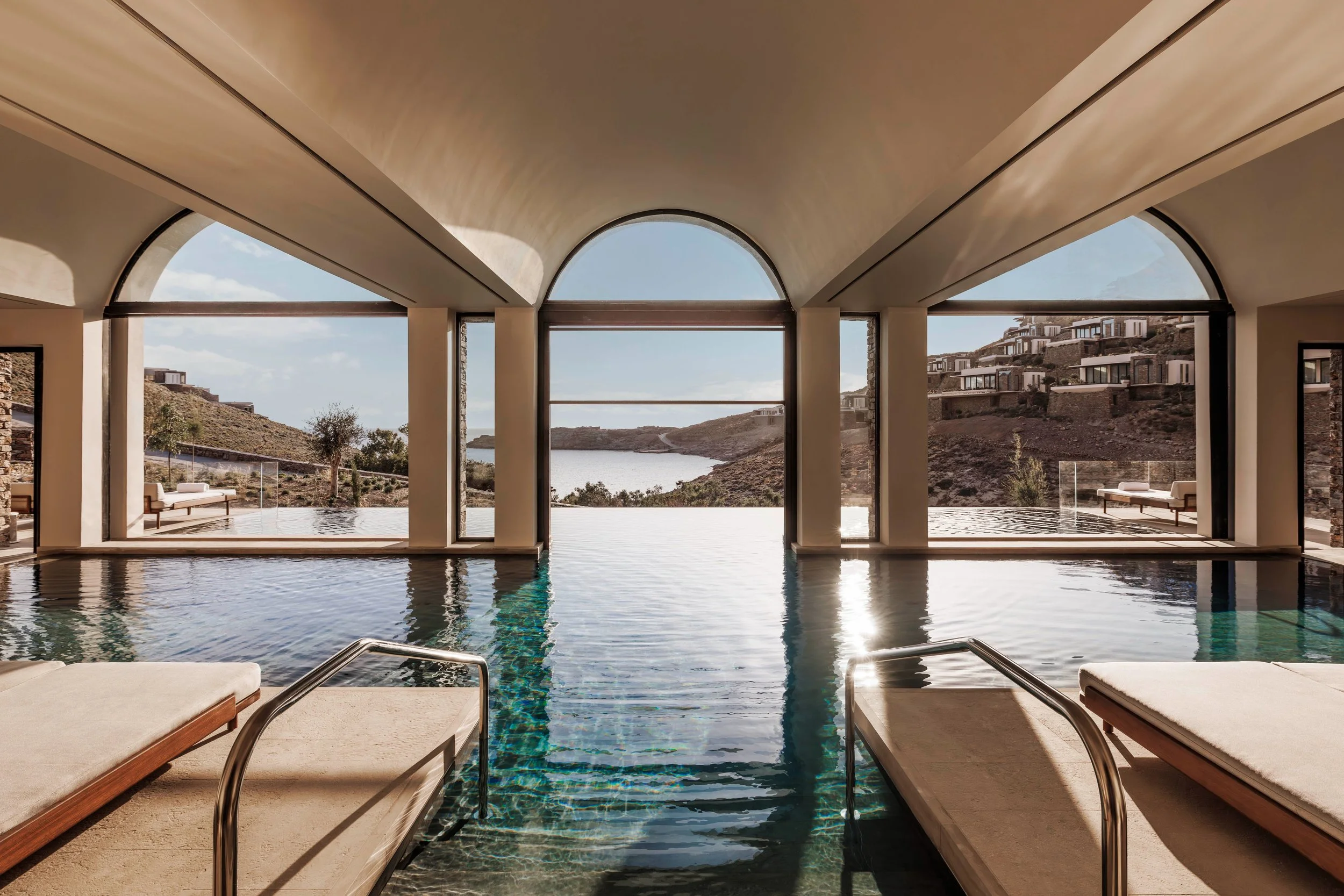 Indoor swimming pool with large floor-to-ceiling windows showing a scenic hillside with modern houses and a body of water in the background.