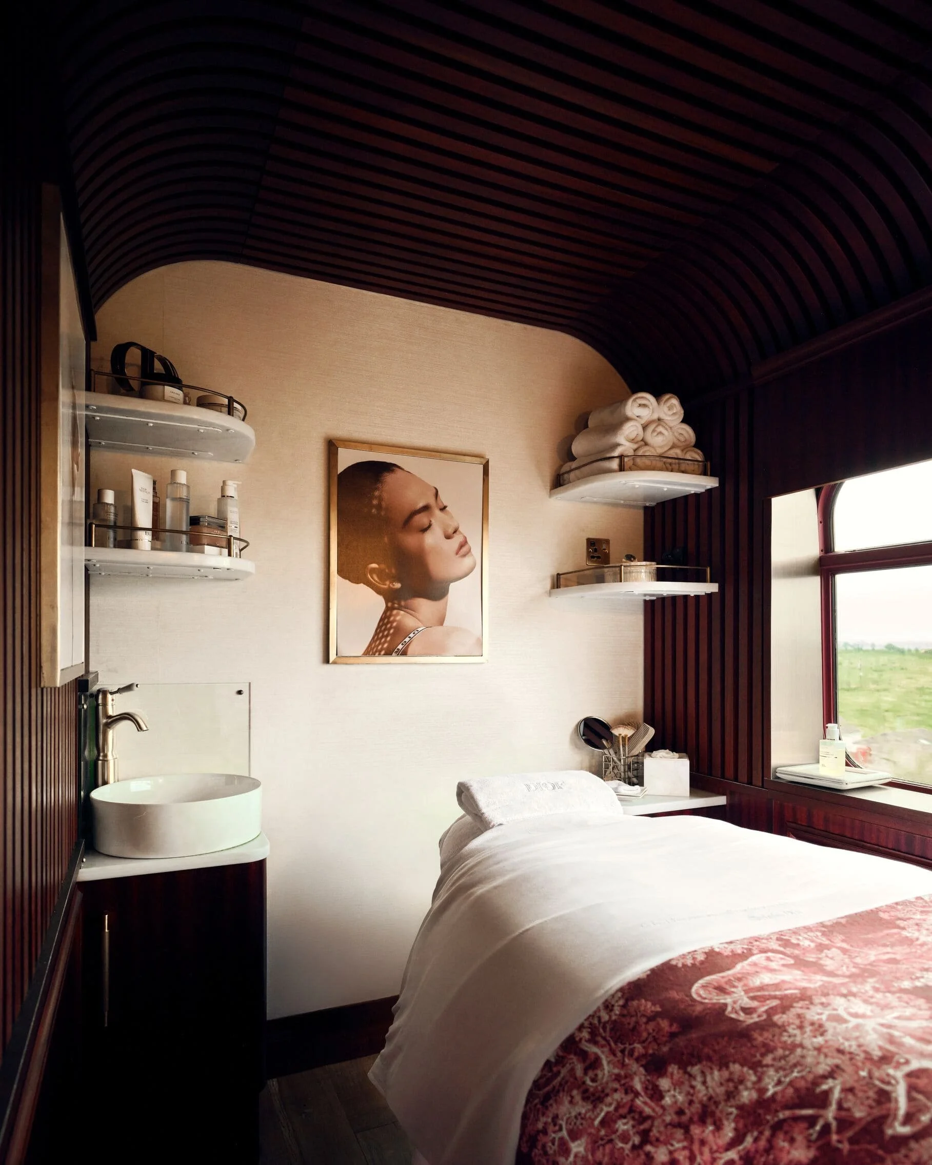 Interior of a spa room with a massage bed, a framed portrait of a woman, shelves with towels and skincare products, and a window showing green fields outside.