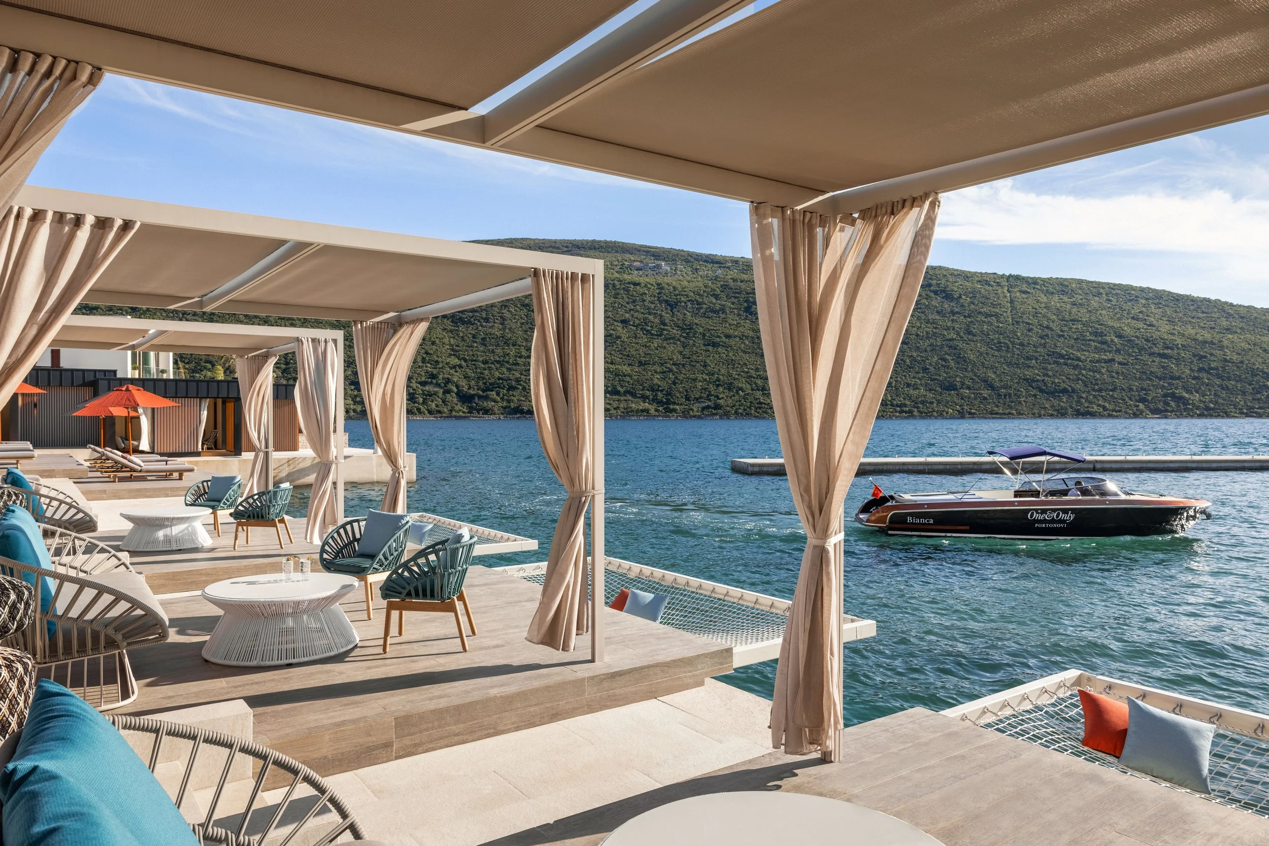 A seaside outdoor lounge area with white tables, cushioned chairs, and curtains under a canopy. In the background, there is water with a boat named Bianca and a mountainous landscape under a blue sky.