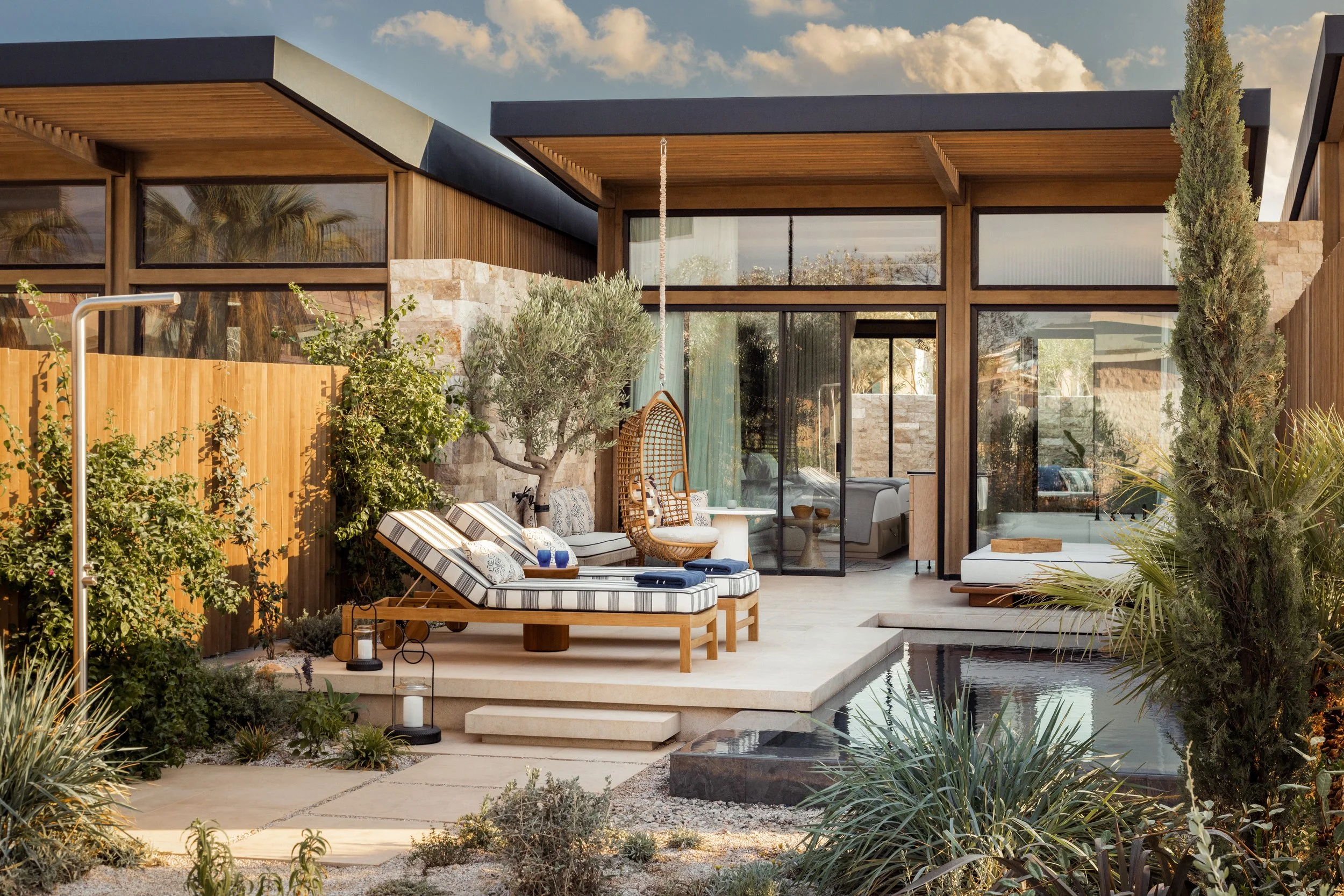 Modern backyard patio with comfortable outdoor furniture, plants, and a pool, adjacent to a contemporary house with large windows and a flat roof. Clear sky with some clouds in the background.