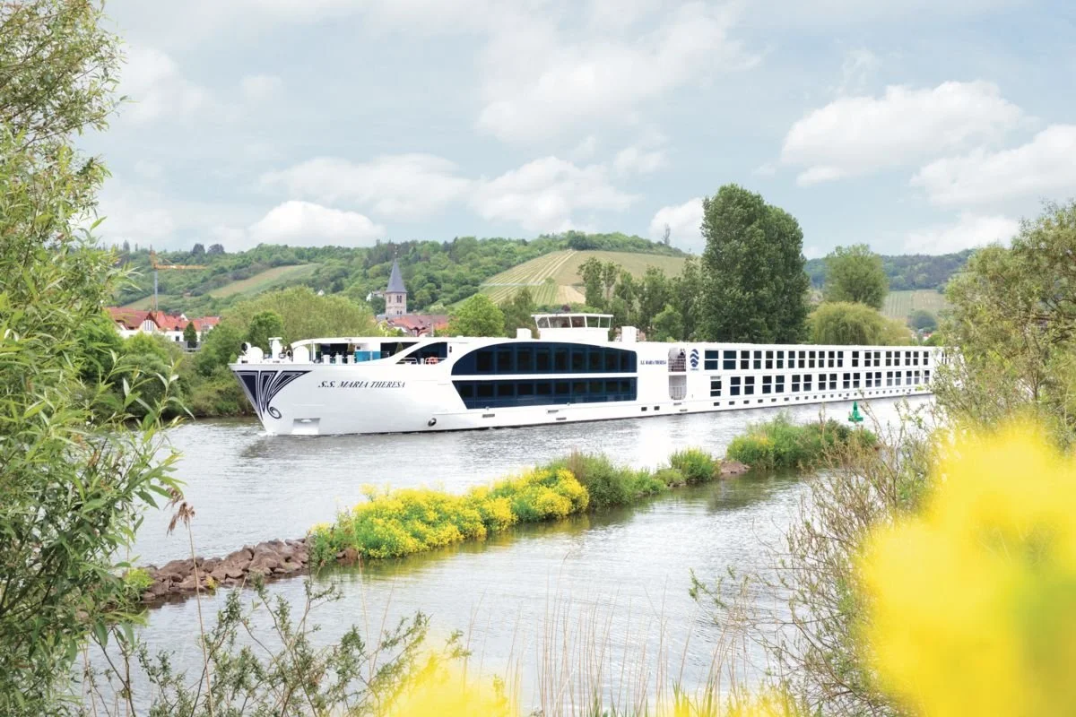 A large white river cruise ship sailing on a river surrounded by green trees and hilly landscape with a village in the background.