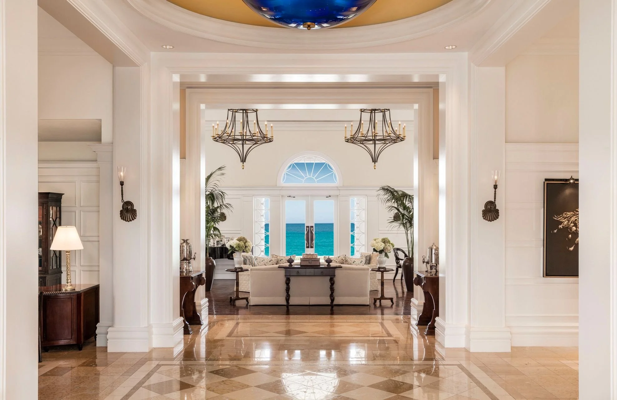 Luxurious living room with white walls, elegant chandeliers, large windows with views of the ocean, and stylish furniture including sofas, tables, and decorative plants.
