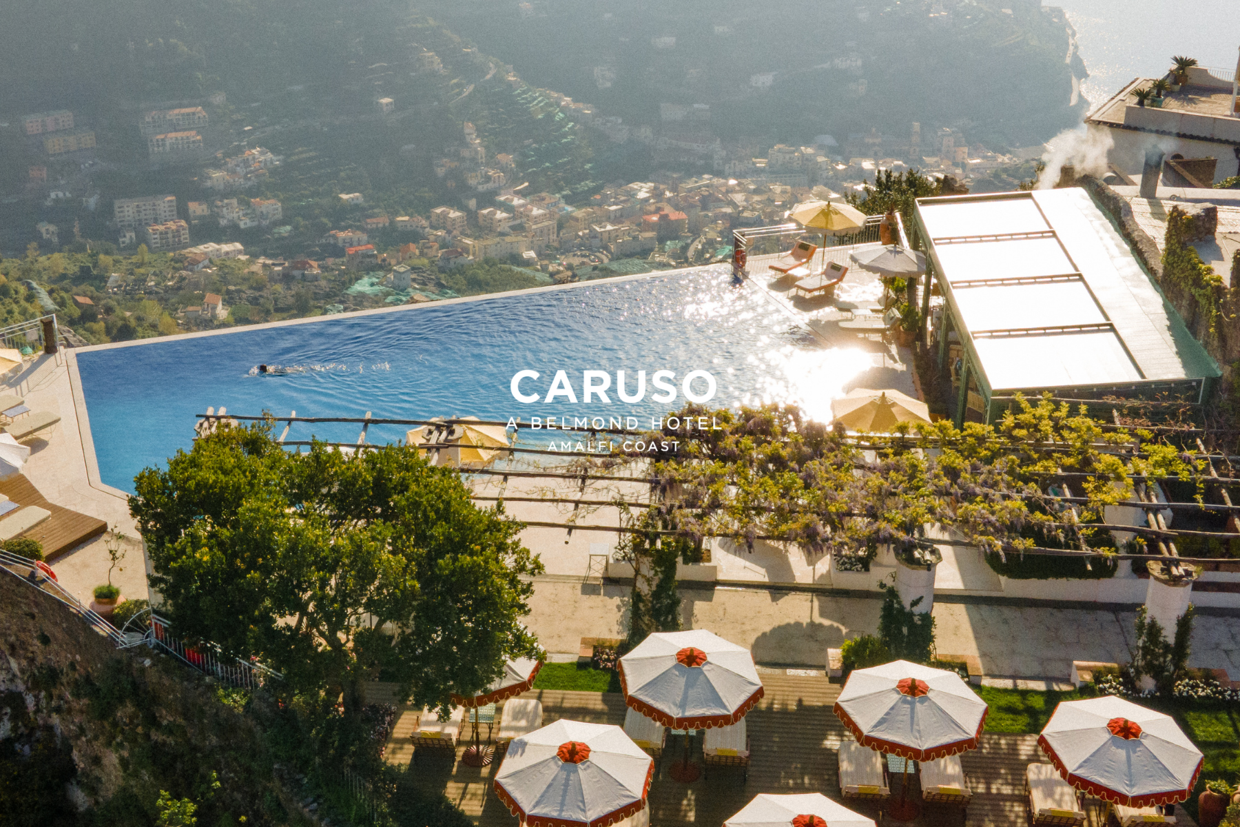 A coastal hotel with an infinity pool, umbrellas, and umbrellas with a scenic view of the mountains, sea, and town below, at Caruso Belmond Hotel on Amalfi Coast.
