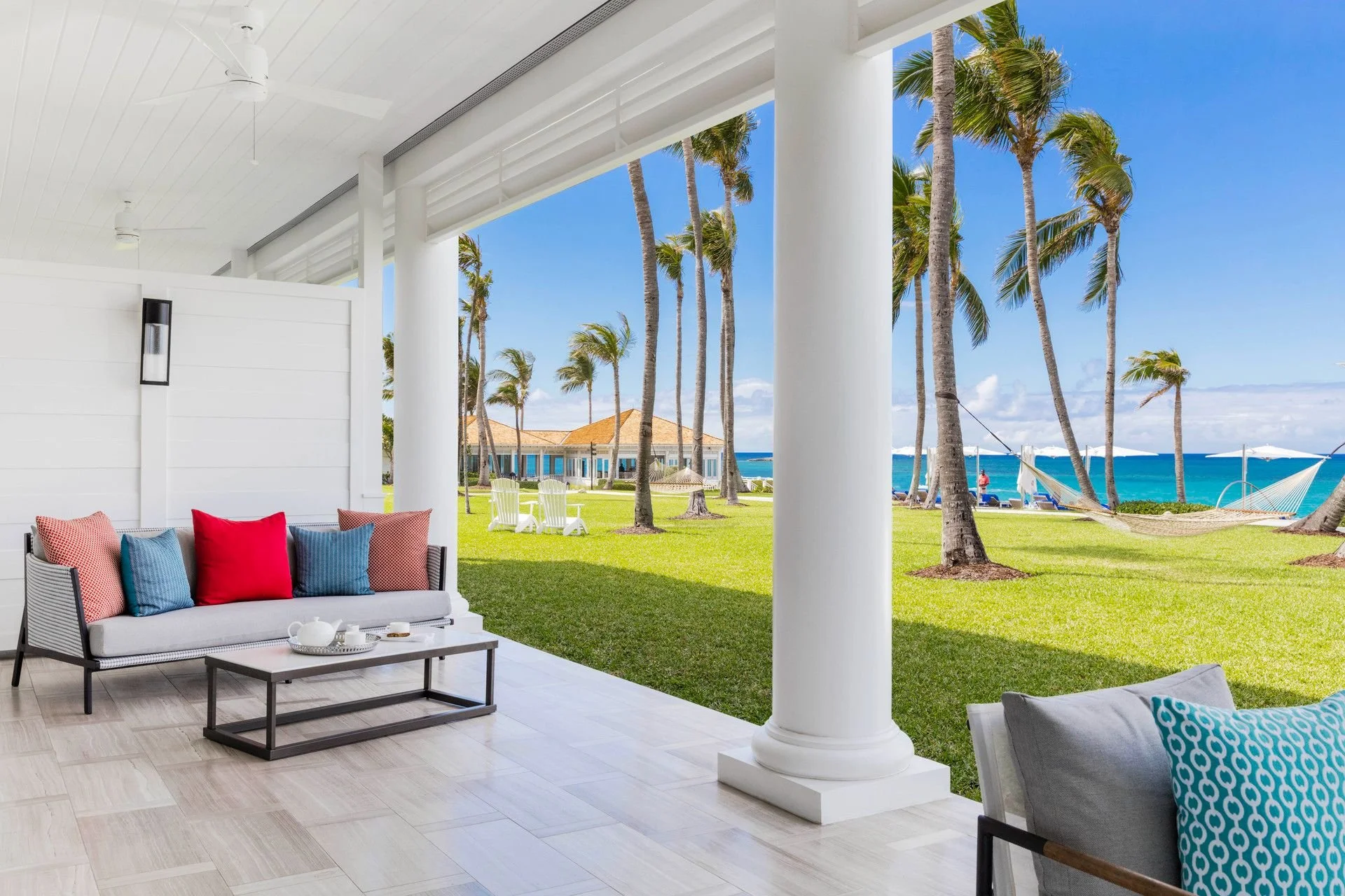 Beachside patio with white columns, furniture with colorful cushions, green grass, palm trees, hammocks, and ocean view.