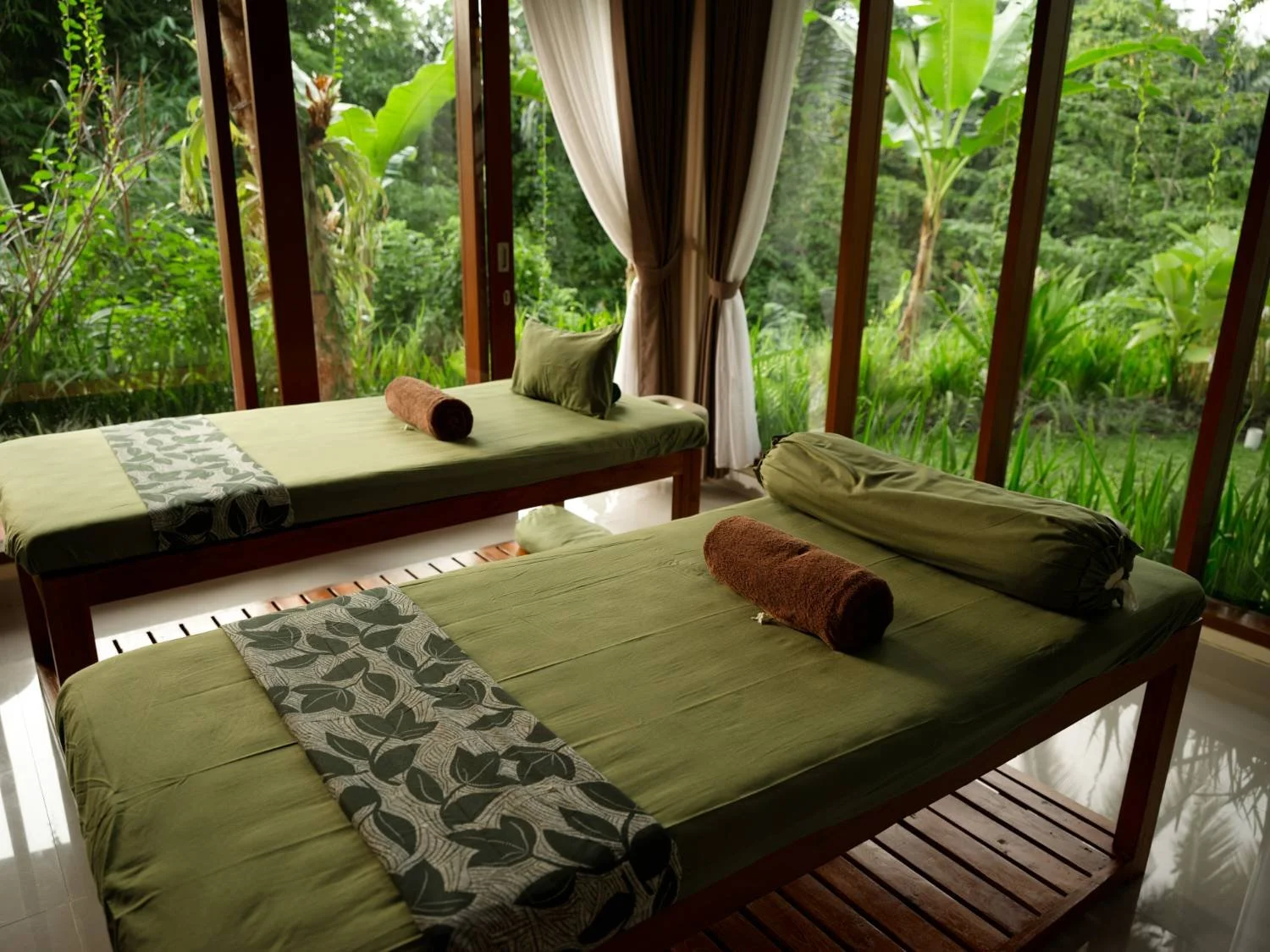 Two massage tables with green sheets and matching pillows, located inside a room with large glass windows showing lush green tropical plants outside.
