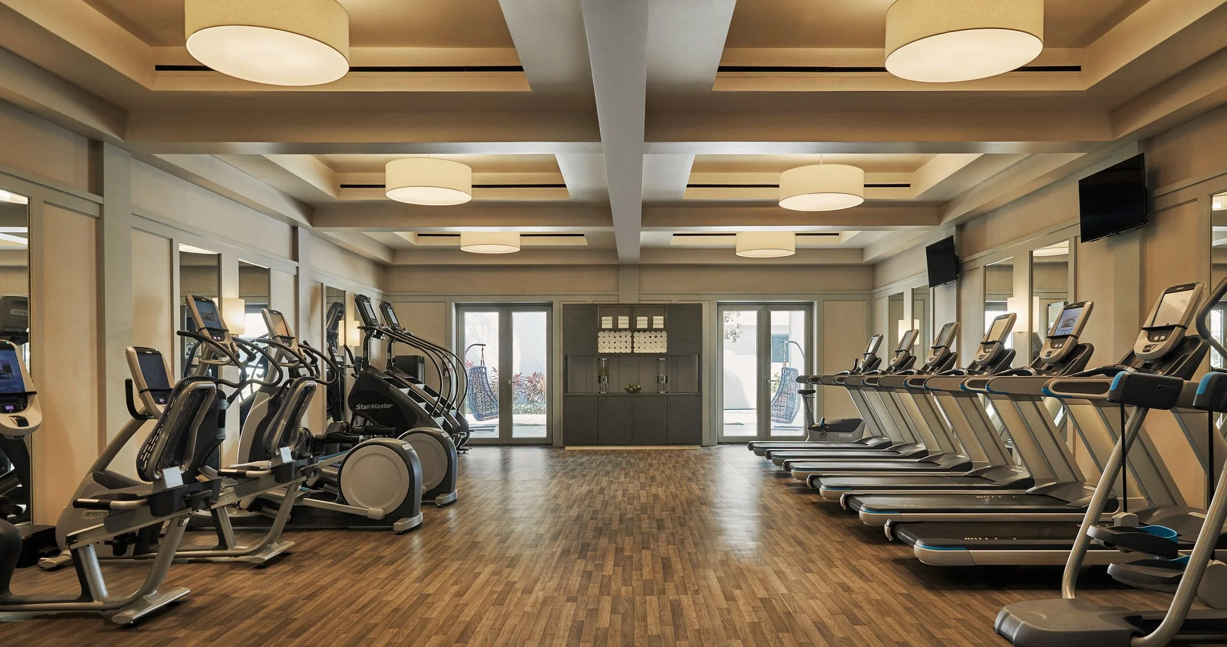 Empty gym with multiple treadmills and elliptical machines, large windows at the back, and ceiling lights.