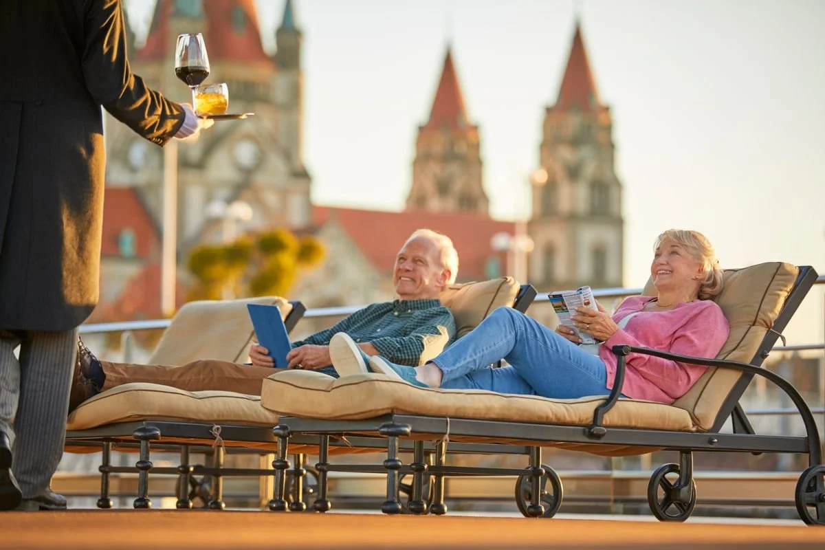 Elderly couple relaxing on lounge chairs on a rooftop patio, smiling at a waiter serving drinks, with a cityscape featuring old-style buildings with red roofs in the background during sunset.