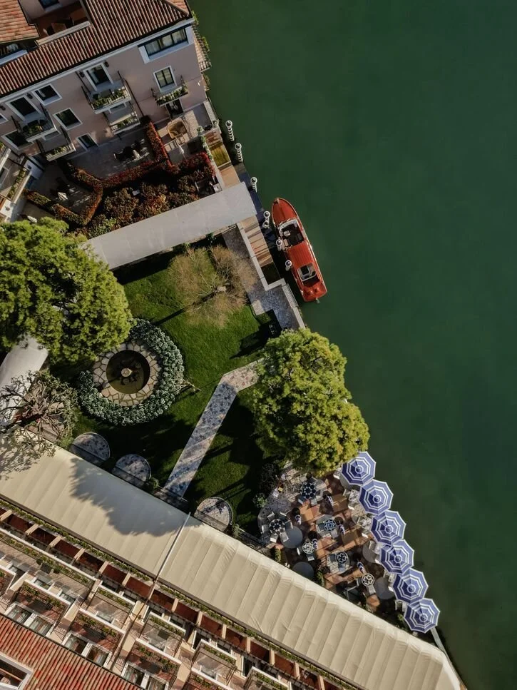 Aerial view of a waterfront residential area with a small lawn, trees, a patio with umbrellas, and a boat docked along the water.