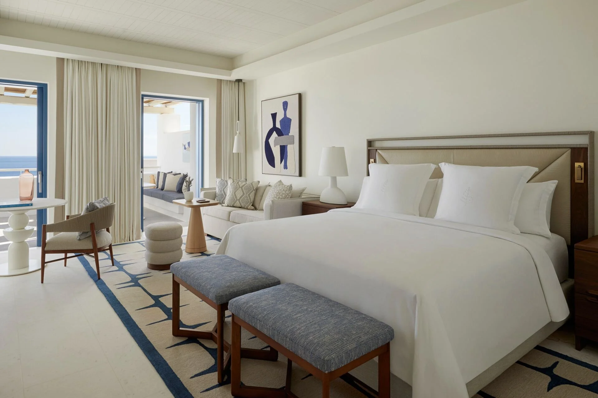 Inside a modern, bright hotel room with white walls and a large bed with white linens. There are two nightstands with lamps, a sitting area with a sofa and decorative pillows, a small table with a vase, a chair, and windowed doors leading to a balcony with ocean views. The room features artwork and a patterned rug.