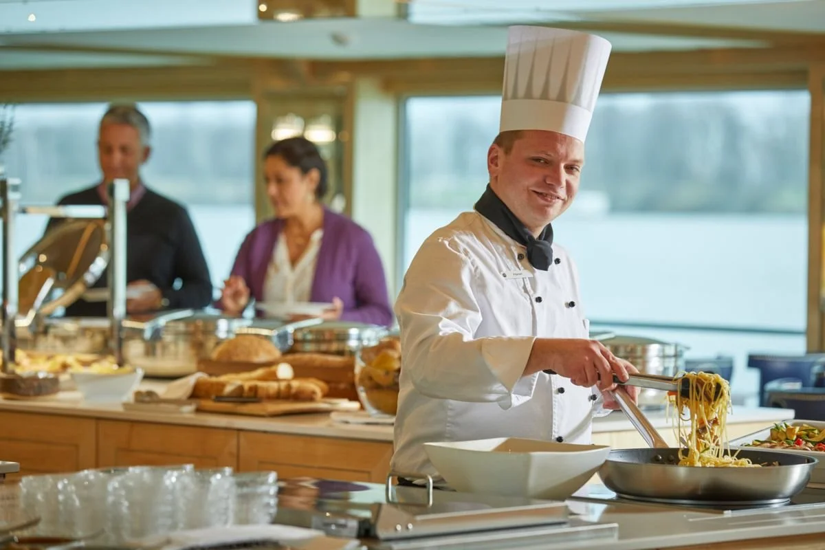 Chef in white uniform and tall chef hat cooking spaghetti in a pan, with people in business attire in the background.