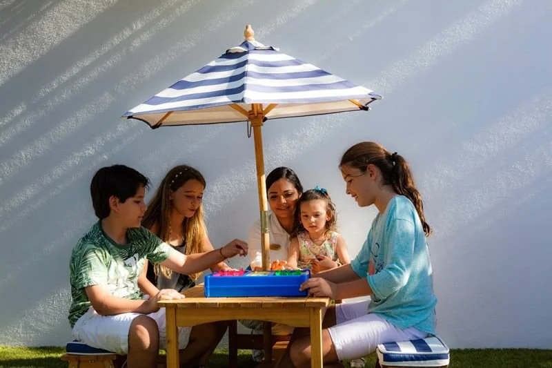 Four children and an adult girl playing a board game under a blue and white striped umbrella outside.