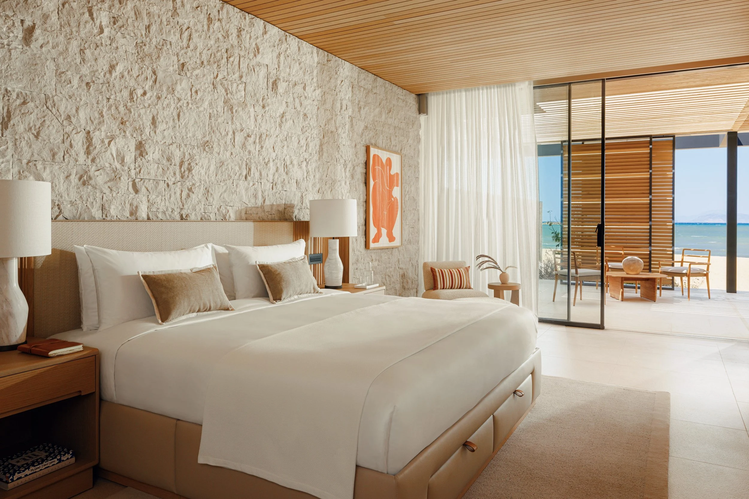 A bedroom with a large bed, beige headboard, white bedding, and pillows. There are bedside tables with lamps and a bookshelf. The room has a textured stone wall, wooden ceiling, and glass sliding doors leading to a balcony with a sea view and outdoor seating.