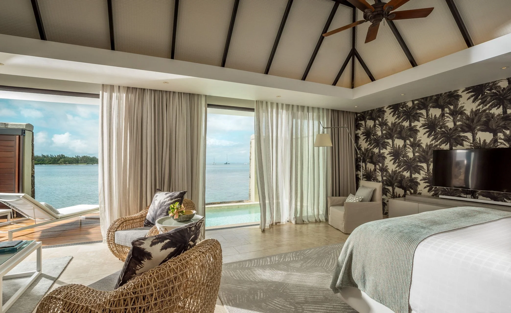 A spacious living room with large windows overlooking water, beach, and sailboats, decorated with palm tree patterned wallpaper, beige curtains, wicker chairs, and modern furniture.