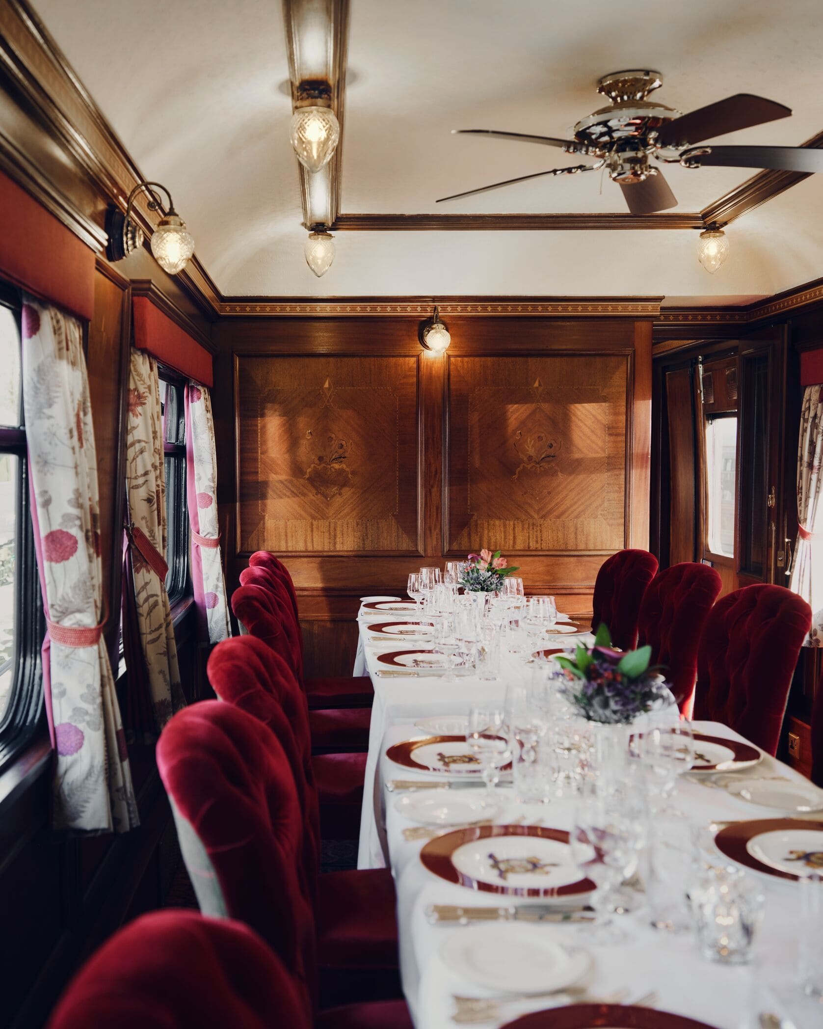 Elegant dining room with a long table covered with a white tablecloth and set with glassware, plates, and cutlery. The room has rich wooden paneling, red velvet chairs, floral curtains, and vintage light fixtures. A ceiling fan is visible.