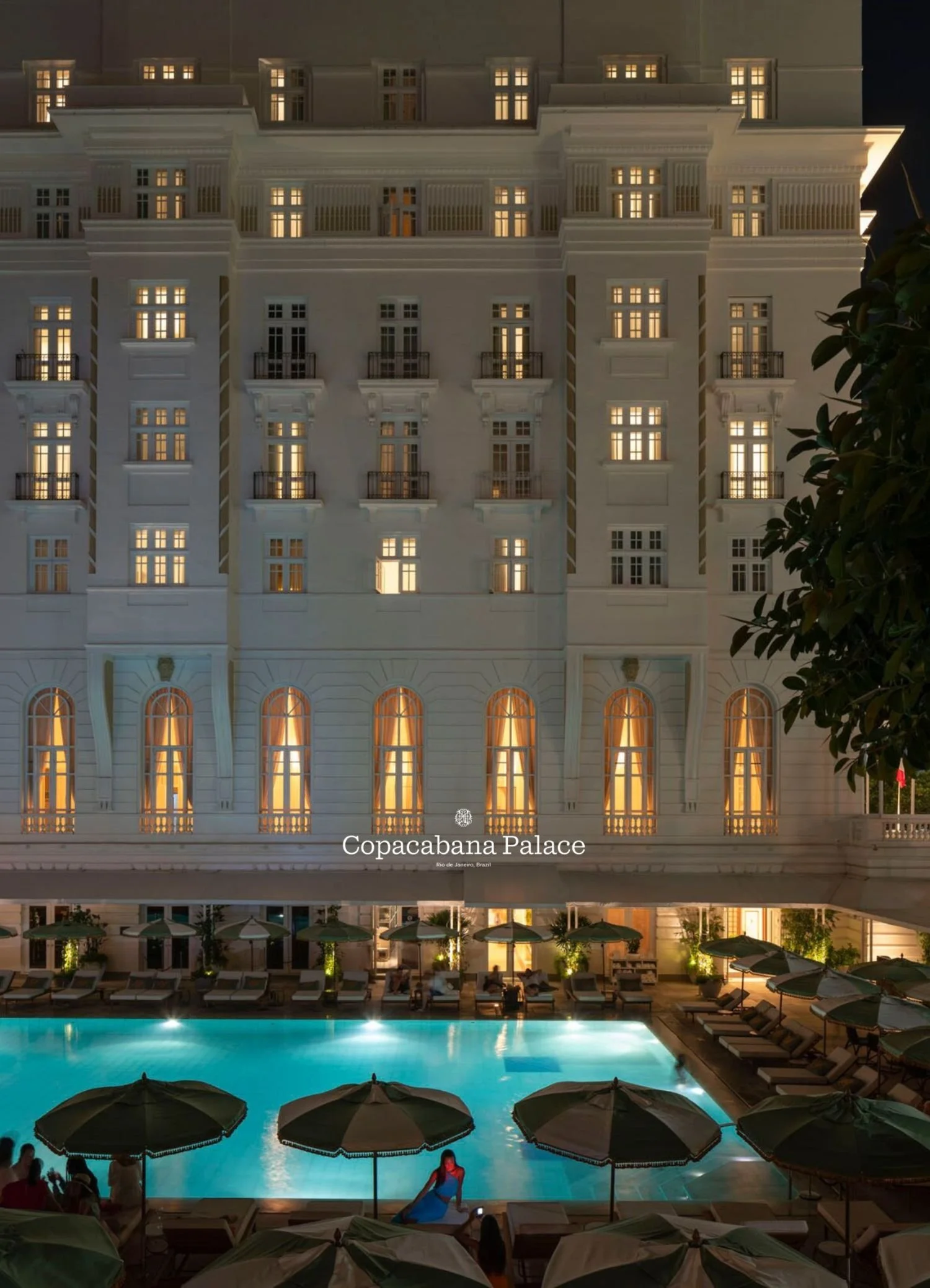 Night view of Copacabana Palace hotel with illuminated windows, outdoor pool area, sun loungers, and umbrellas under a dark sky.