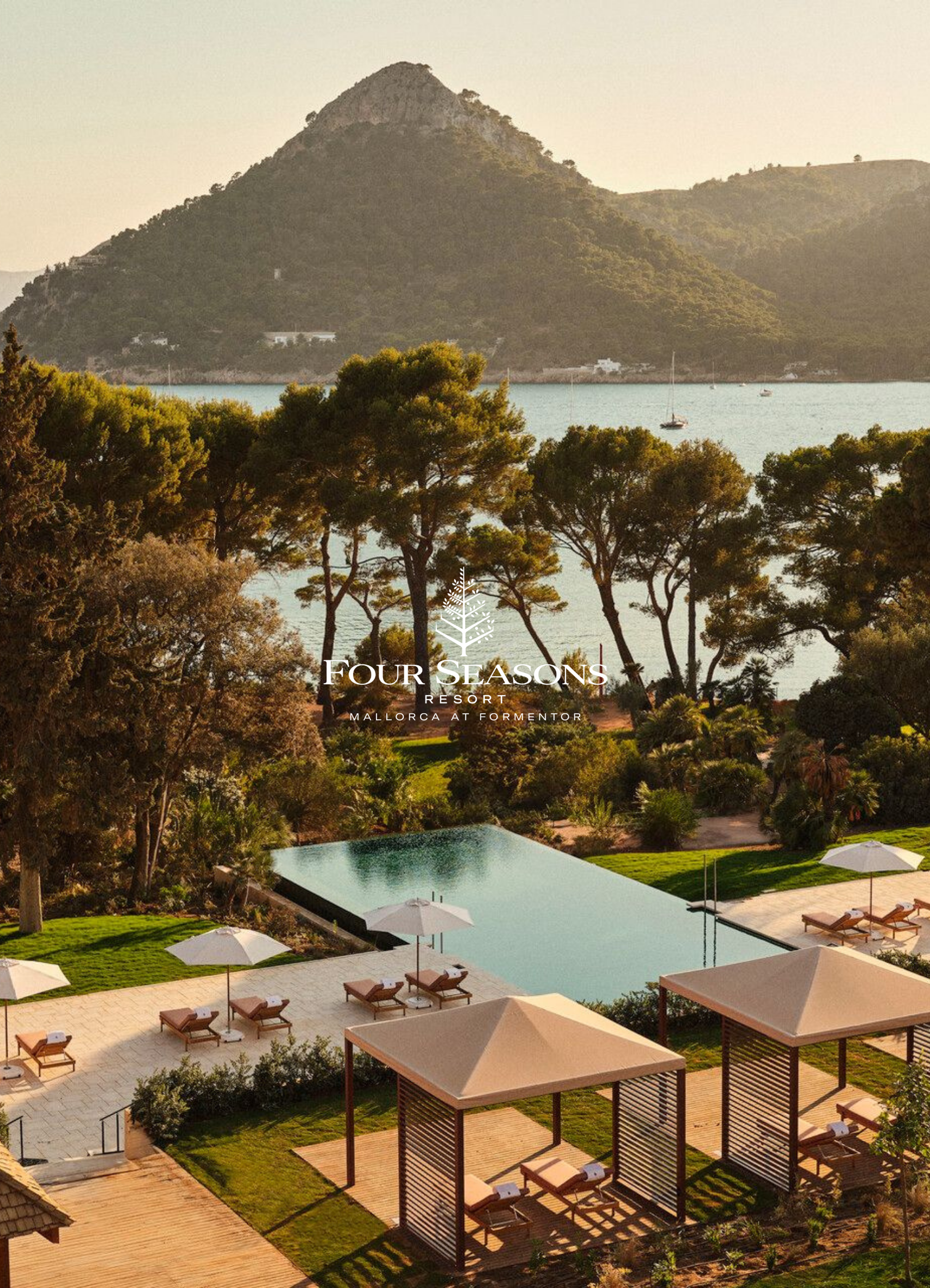 A scenic view of Four Seasons Resort Mallorca at Formentor, showing a mountain in the background, a body of water with boats in the middle, and a pool area with lounge chairs and umbrellas in the foreground.