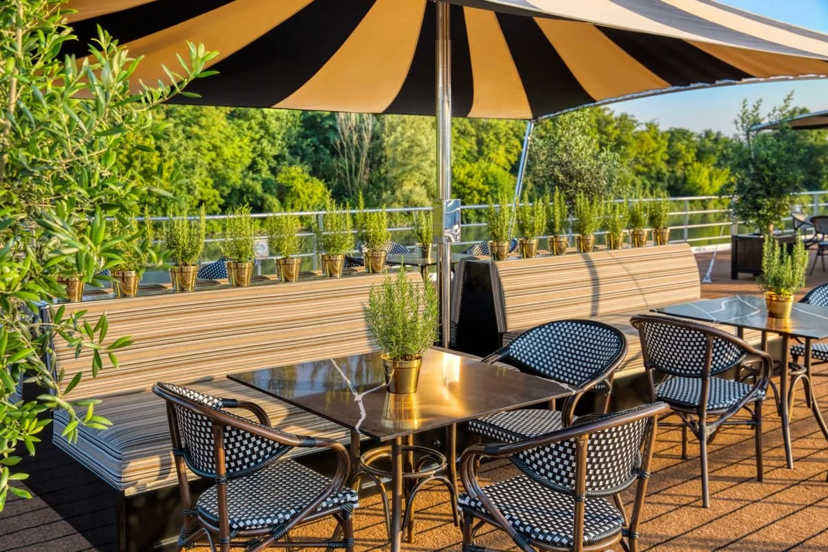 Outdoor patio with potted herbs, black and white checkered chairs, and a large striped umbrella, overlooking green trees.