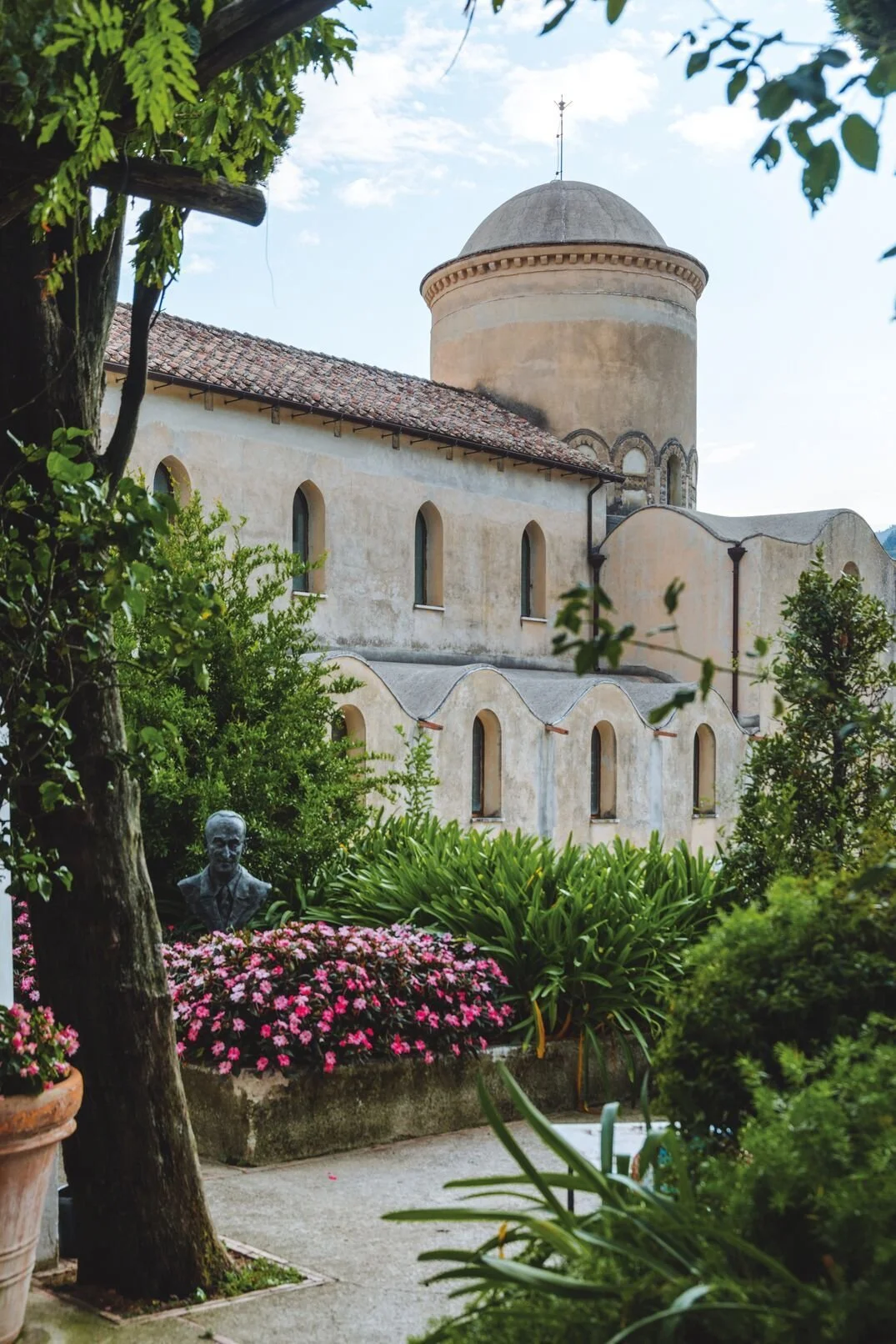 A historic church with a round tower, arched windows, and a tiled roof, surrounded by lush green plants, flowers, and trees, with a small bust sculpture visible among the foliage.