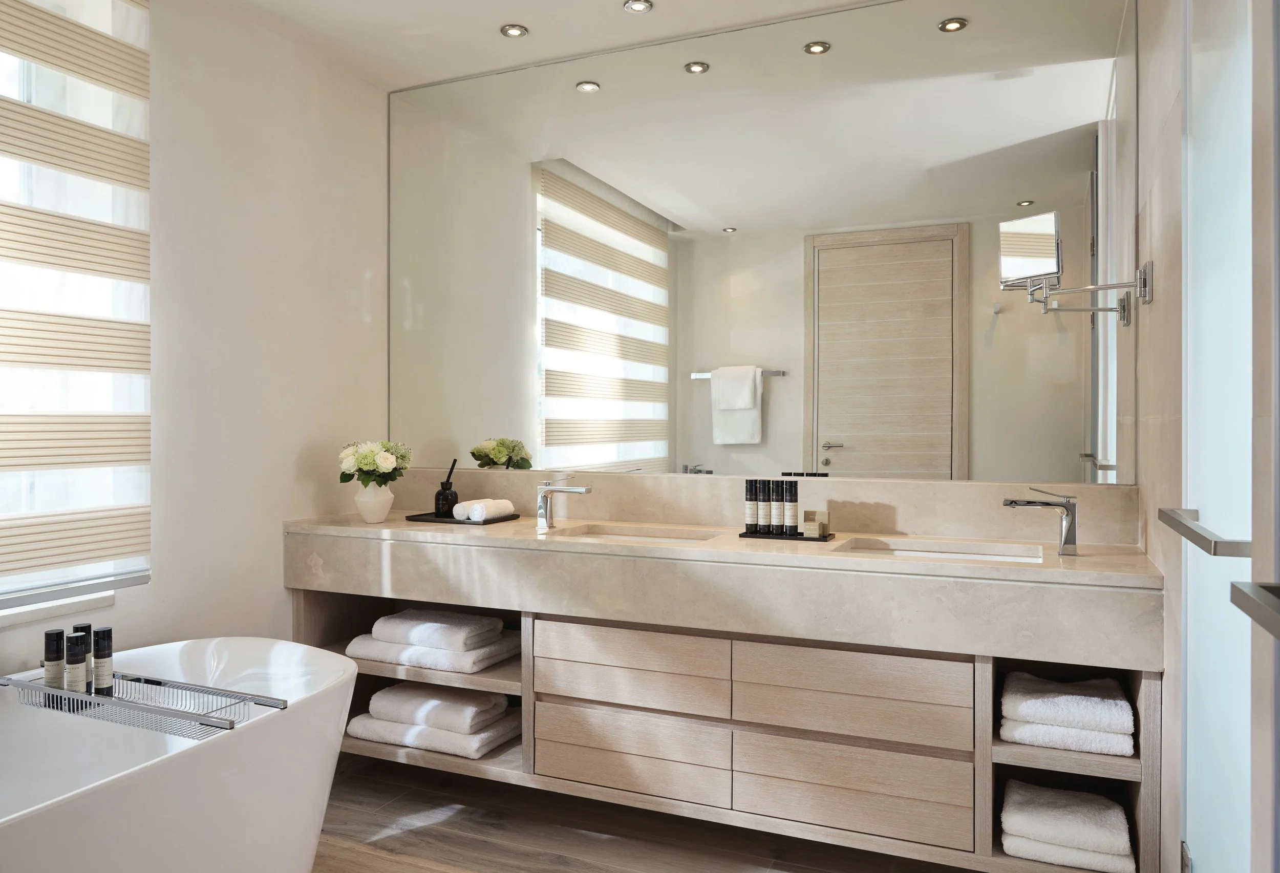 A modern bathroom with a large mirror above a beige marble countertop. The countertop has toiletries, towels, and decorative flowers. There is a white bathtub in the foreground with bottles of bath products. The bathroom features light-colored wood cabinets and a window with horizontal blinds.