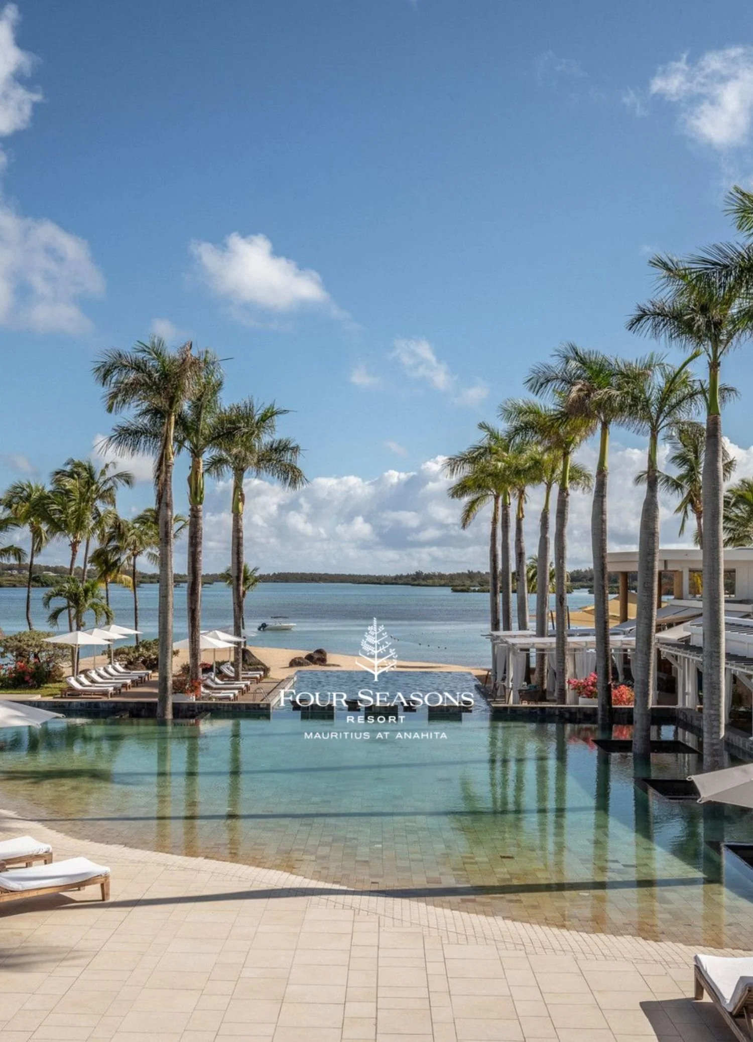 View of a luxurious tropical resort with a swimming pool, palm trees, lounge chairs, umbrellas, and a view of the ocean in the background under a partly cloudy sky. The resort is called Four Seasons Resort Mauritius at Anahita.