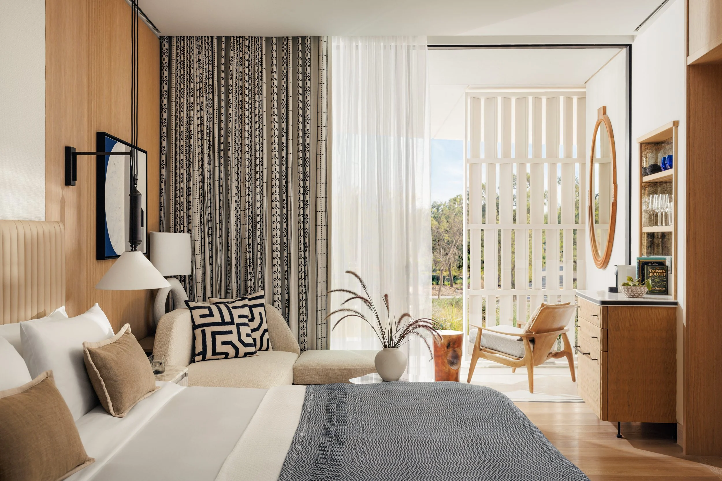 Modern bedroom with a large bed, beige pillows, and a beige headboard. There's a sitting area with patterned pillows, a white sofa, a lamp, and a decorative plant. Large windows with patterned curtains and sheer drapes reveal an outdoor view.