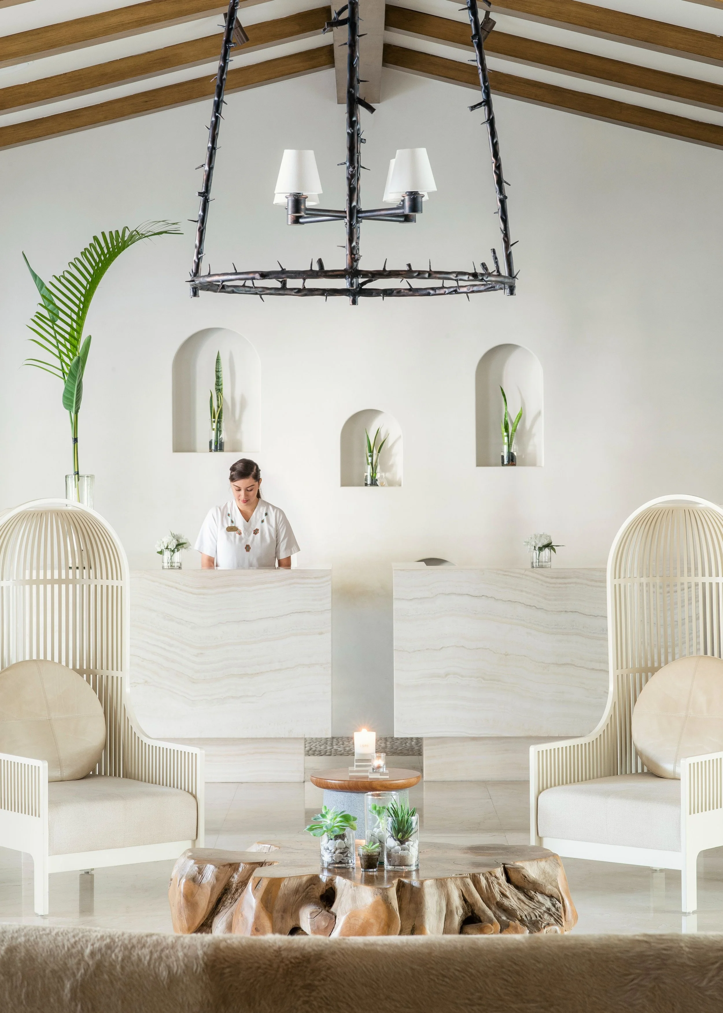 A minimalist, modern interior with a white reception desk, behind which a woman in a white uniform is working. The background features three arched niches with green potted plants, white walls, and a large black chandelier with white lampshades hanging from a wooden-beamed ceiling. In the foreground, a rustic wooden table holds three glass vases with succulents and stones, and a lit candle.