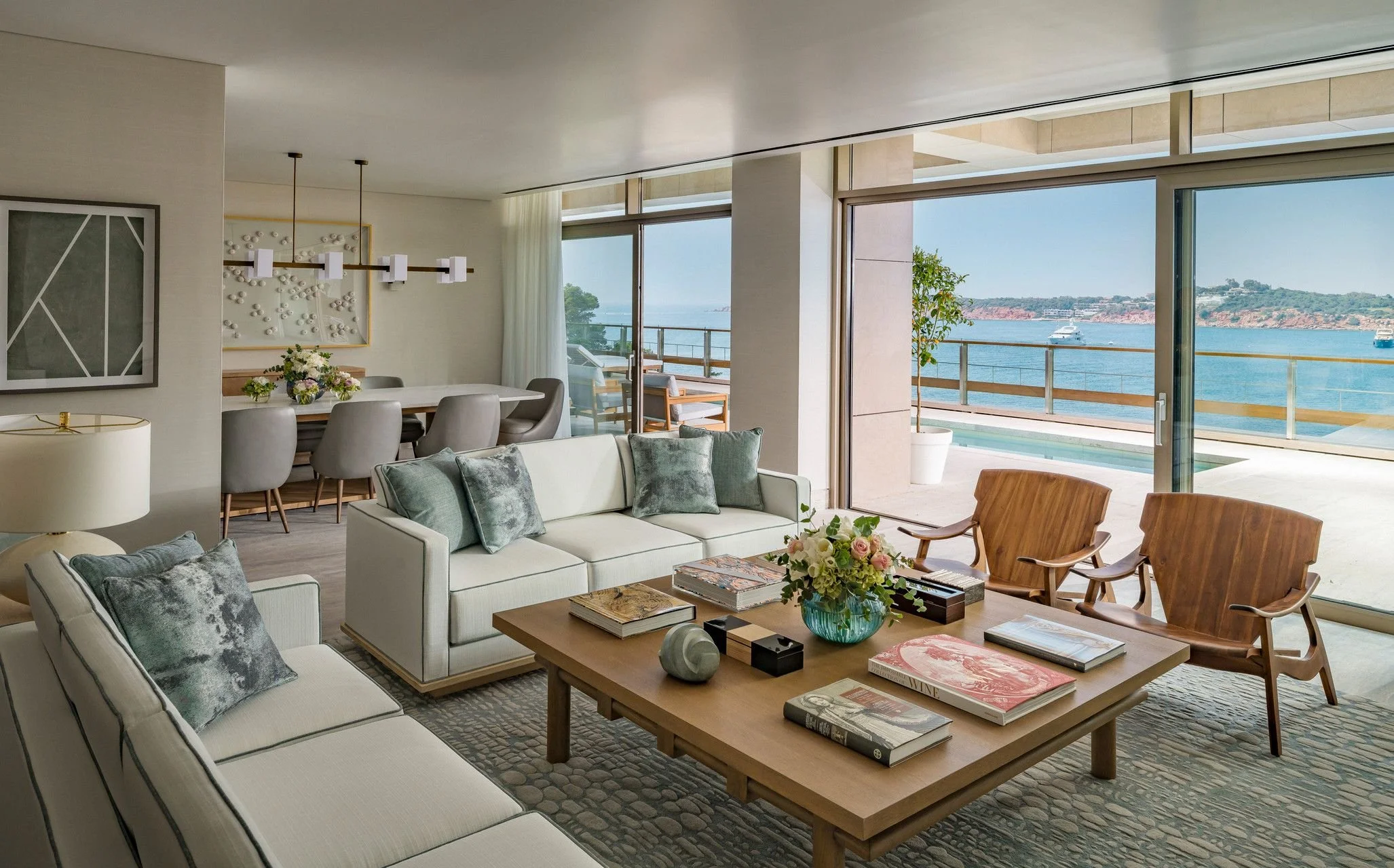 Living room with white sofas, gray pillows, wooden coffee table with books and flowers, floor-to-ceiling glass doors showing waterfront view with boats, and modern art on the walls.