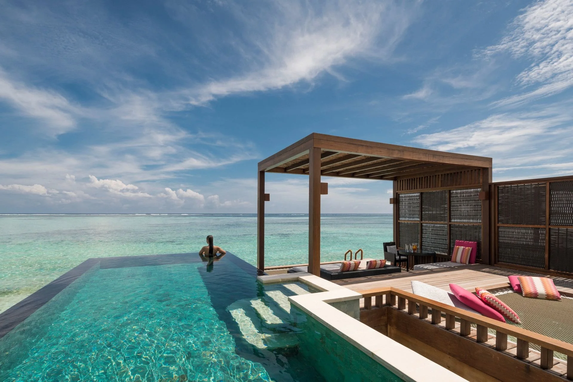 A woman relaxing in an infinity pool overlooking the ocean at a beachside resort, with a wooden cabana and outdoor seating area under a partly cloudy sky.