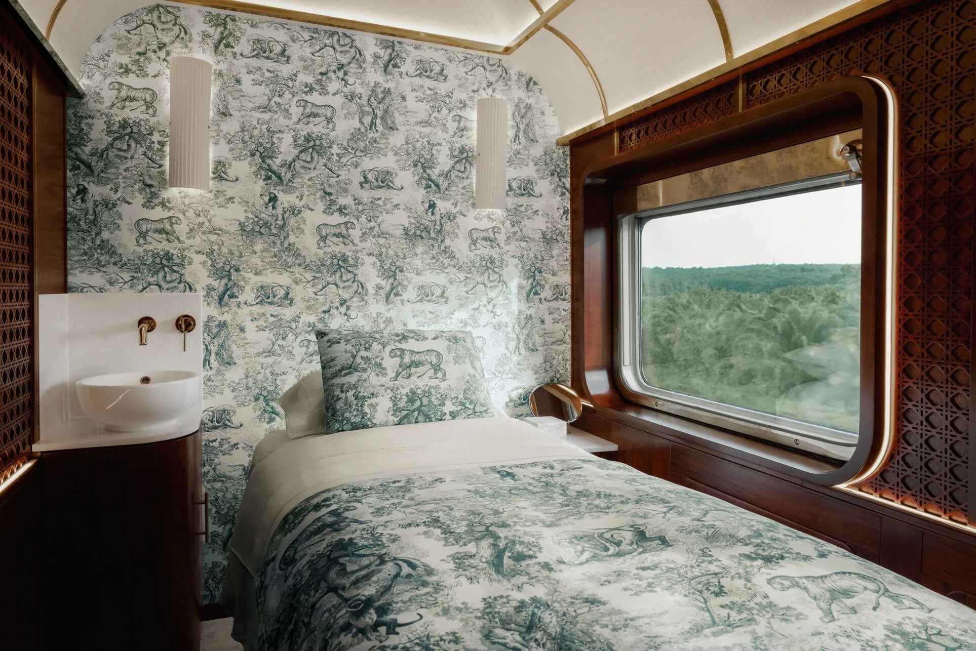 Interior of a train compartment with a bed, a large window showing a green landscape, a small sink, and two hanging lights. The walls have a snake and tree patterned wallpaper, and the room features wood accents.