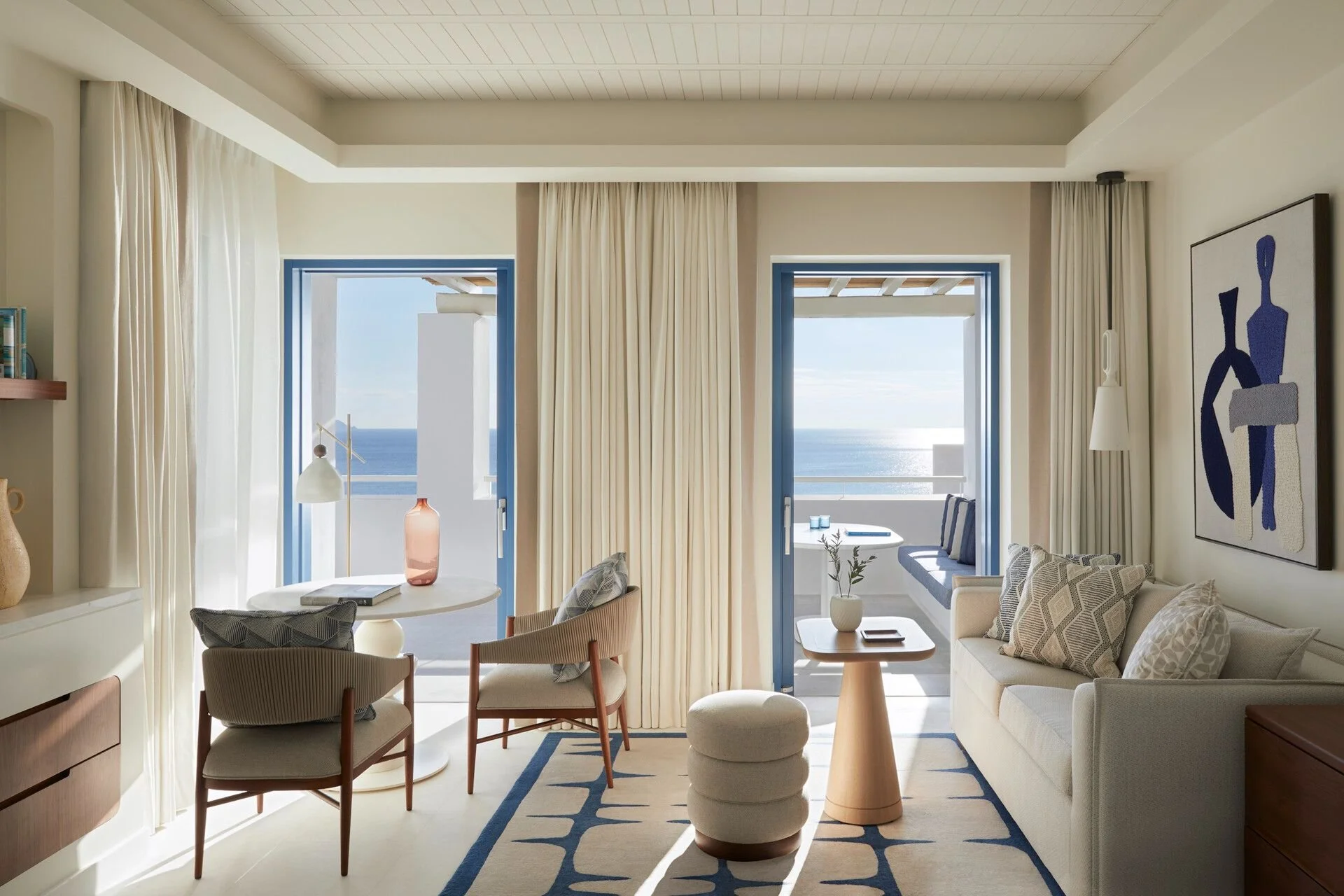 Living room with large sliding glass doors opening to a balcony with ocean view, white walls, beige curtains, modern furniture, abstract wall art, and natural light.