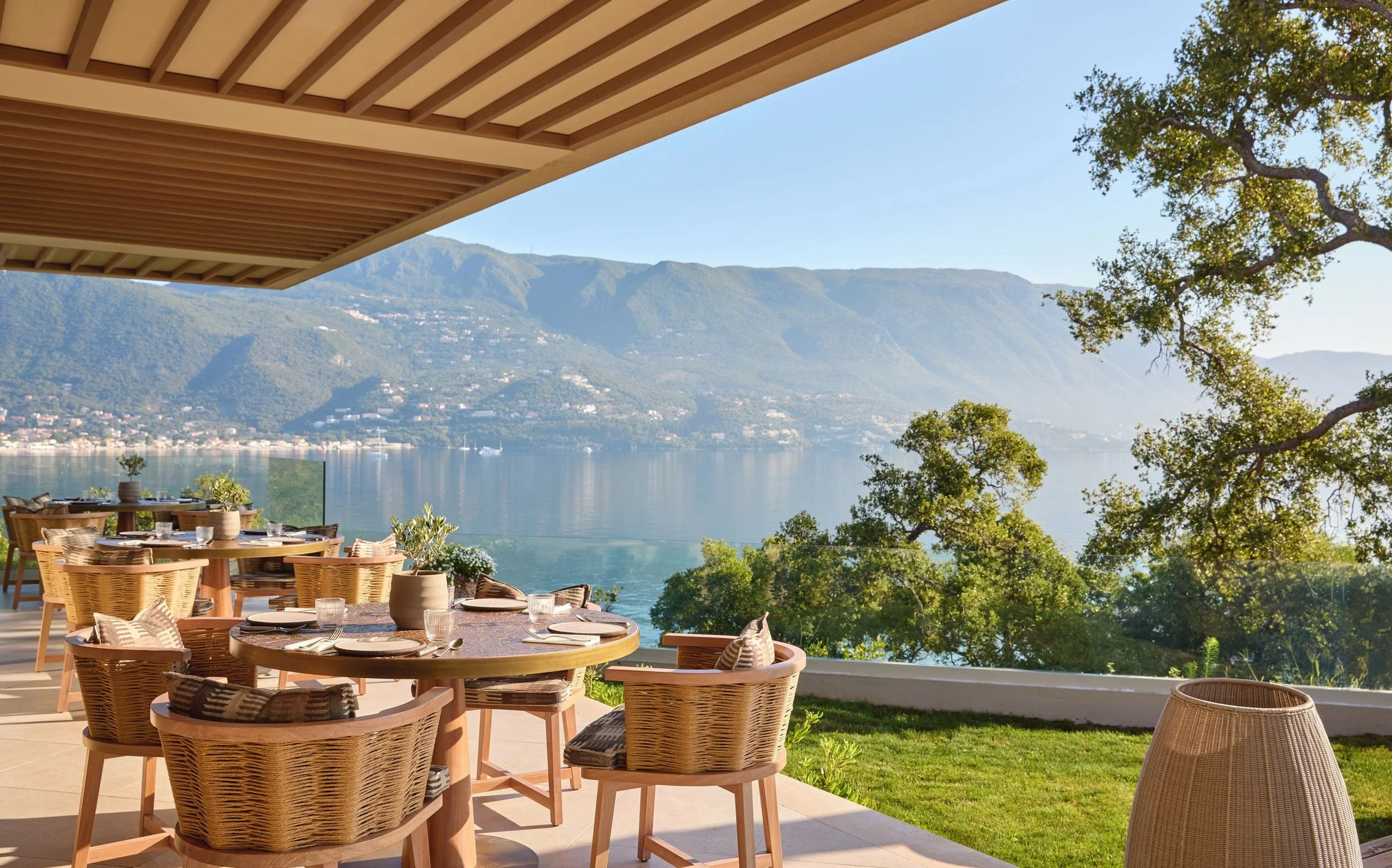 Outdoor dining area overlooking a lake with mountains in the background, partially shaded by a modern overhang, with wicker chairs and round tables set for a meal.