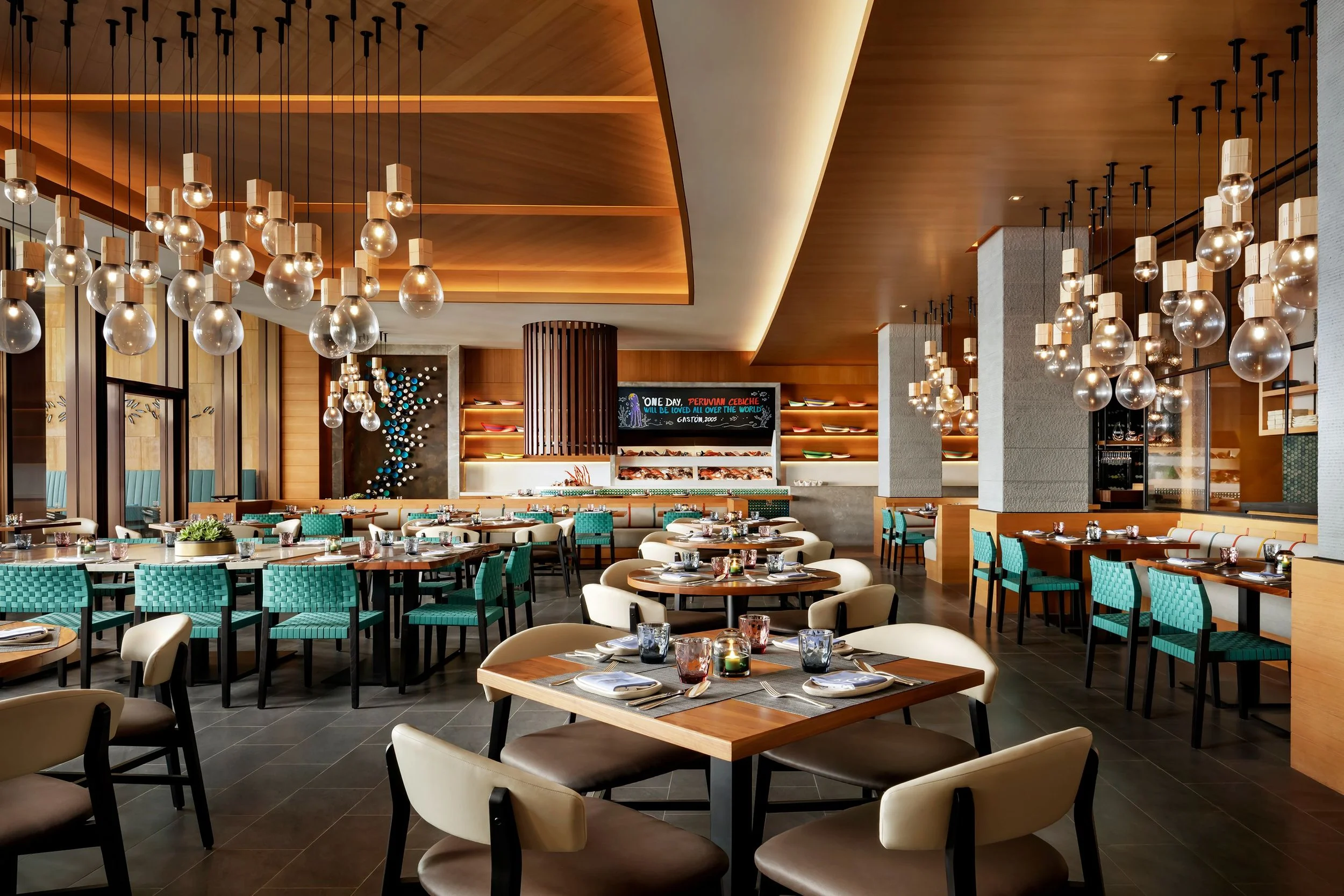 Modern restaurant interior with hanging glass pendant lights, round tables set with glassware and cutlery, and teal and beige chairs. Wood-paneled ceiling and walls create a warm ambiance.