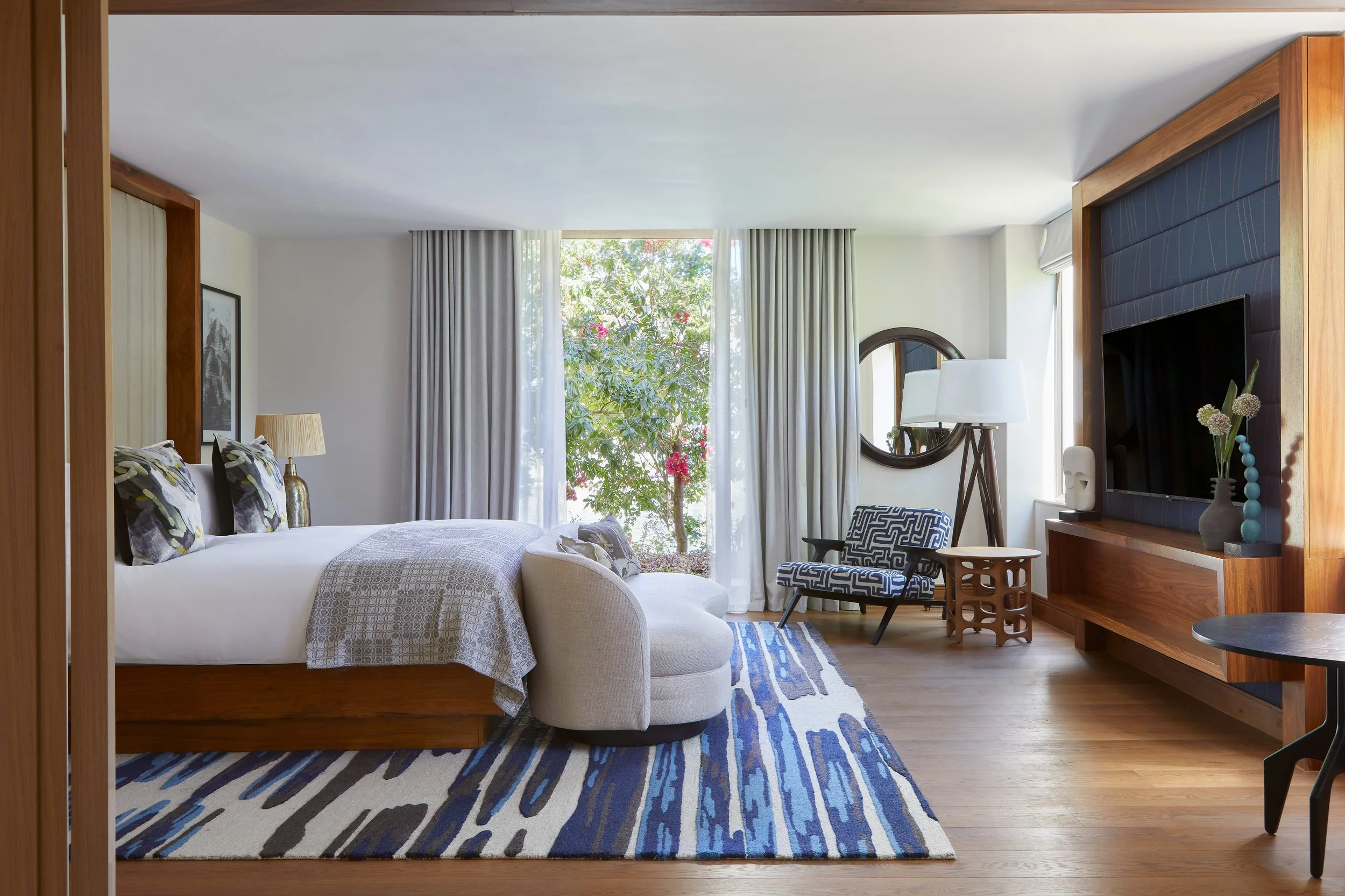A living room with a bed on the left, a window with curtains in the center, and a TV on the right, decorated with vases and artwork.