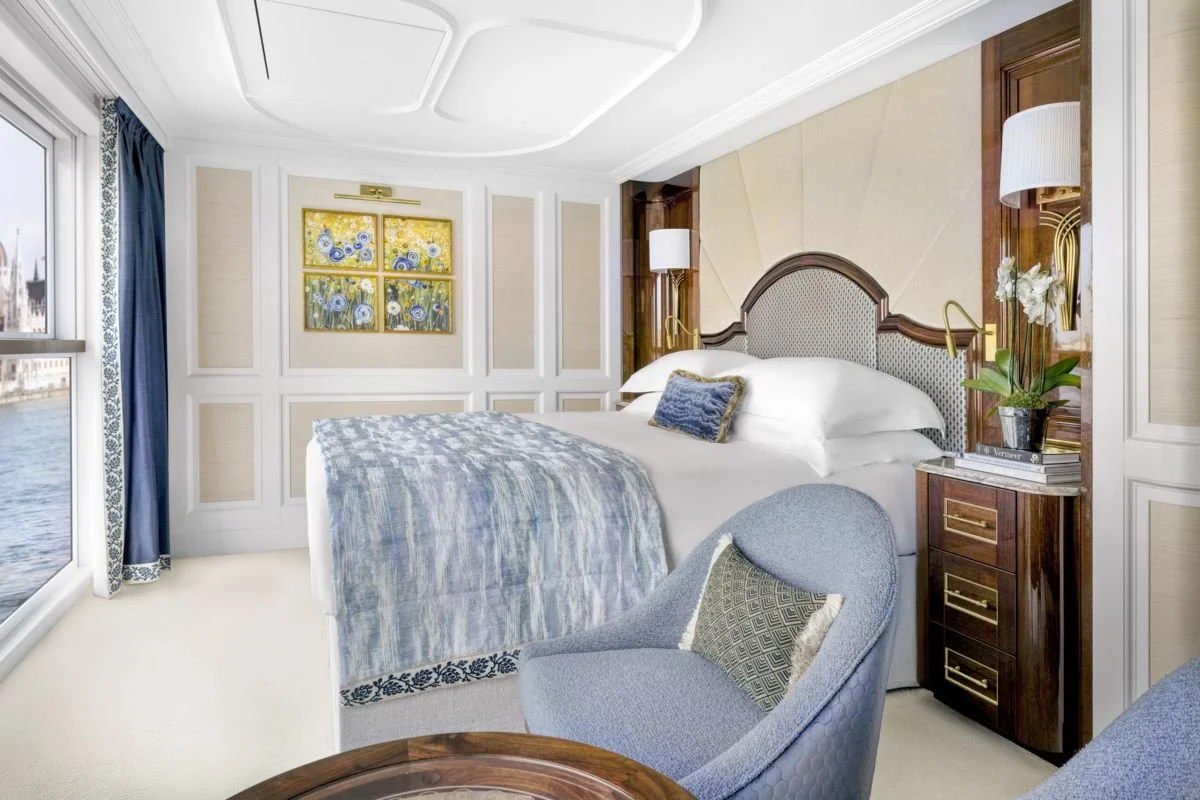 A luxurious bedroom with a large bed, blue and beige-themed decor, a nightstand with books and flowers, a blue armchair, a round wooden table, a window with a river view, and wall art featuring Vincent van Gogh's sunflower painting.