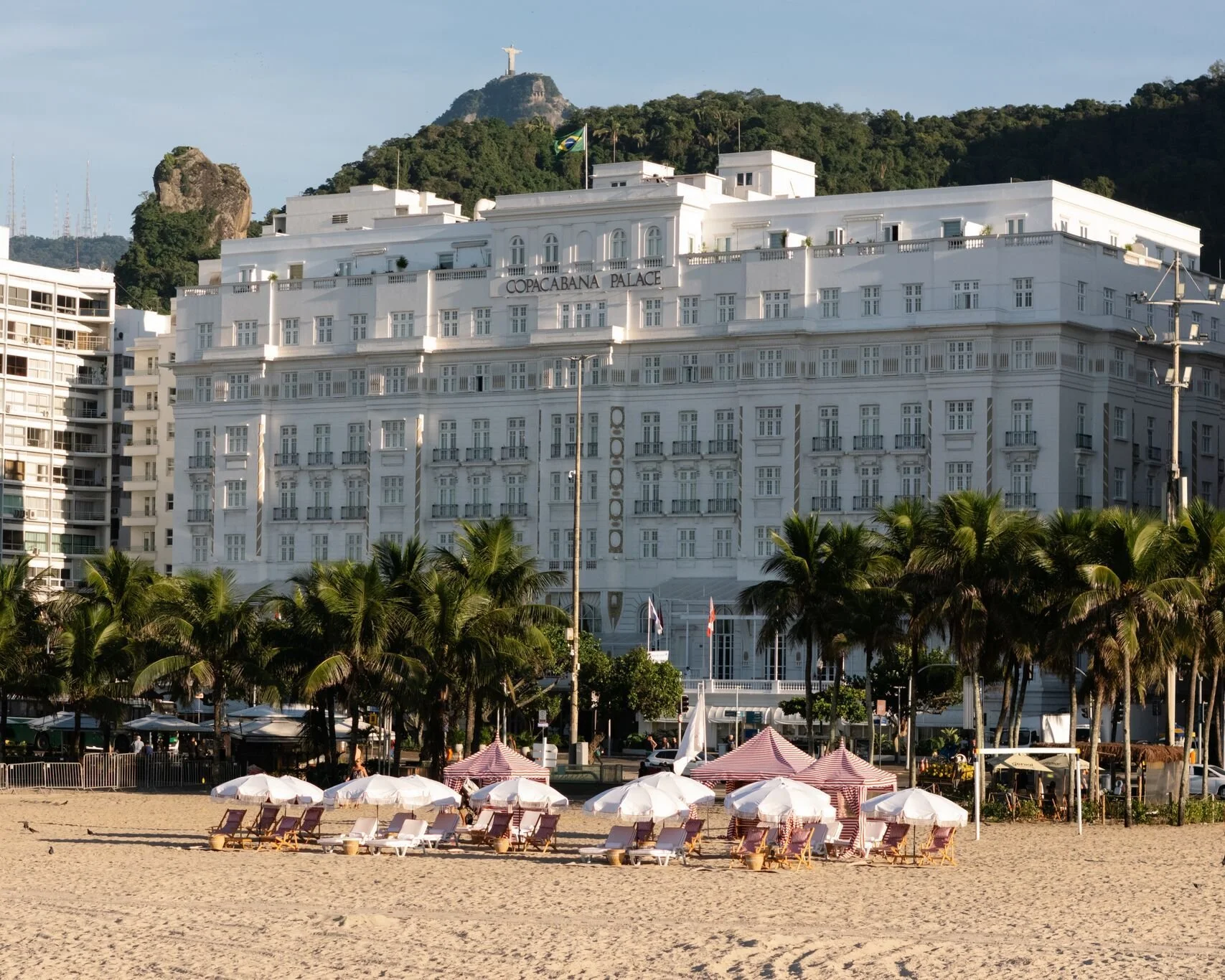 Beach with rows of white lounge chairs and umbrellas, palm trees, and a large white hotel building labeled 'Copacabana Palace' in Rio de Janeiro, Brazil, with mountains and Christ the Redeemer statue in the background.