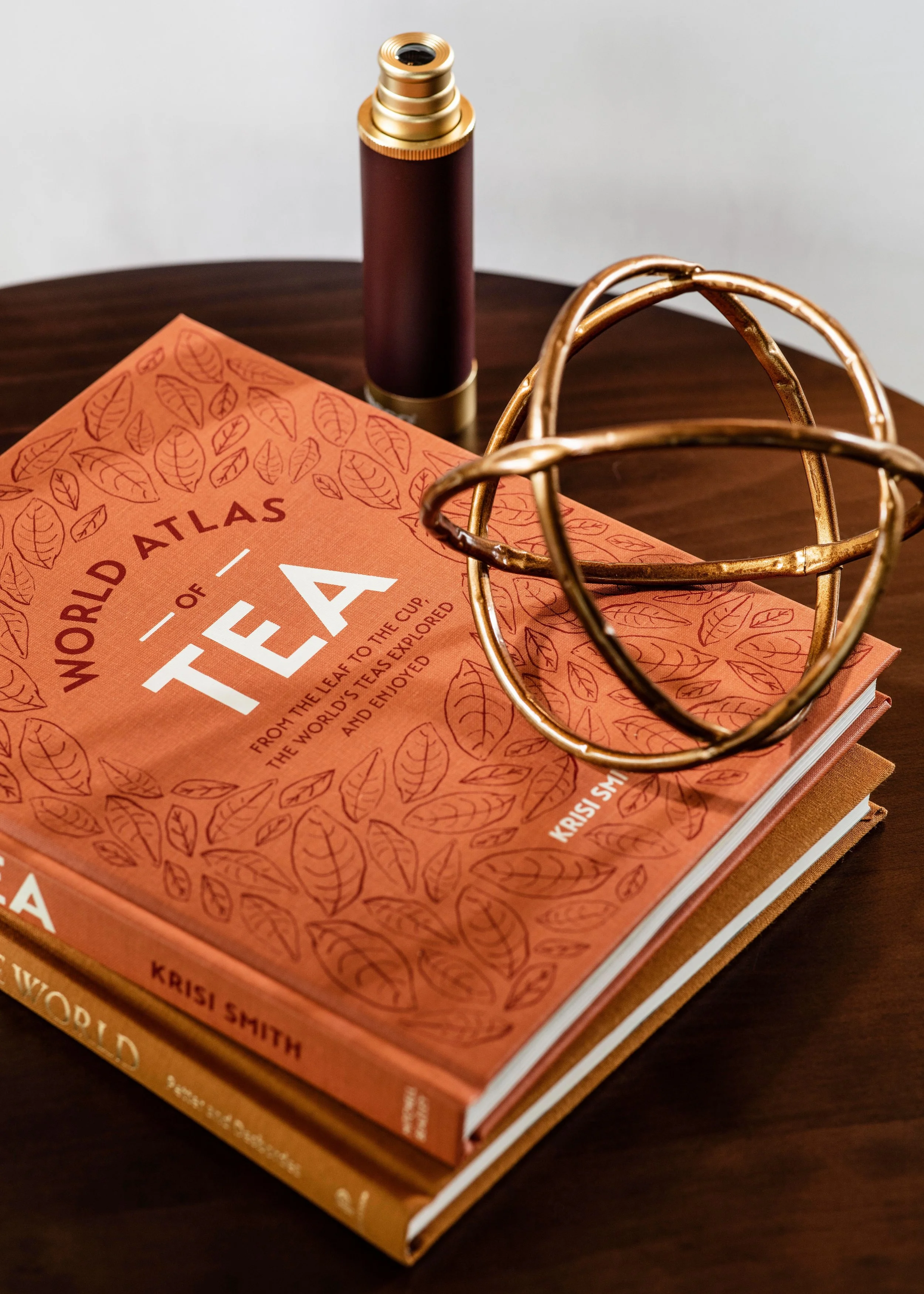An orange book titled "World Atlas of Tea" resting on a dark wooden surface, with a metallic gold geometric sculpture and a metallic tube with a ribbed design standing upright.