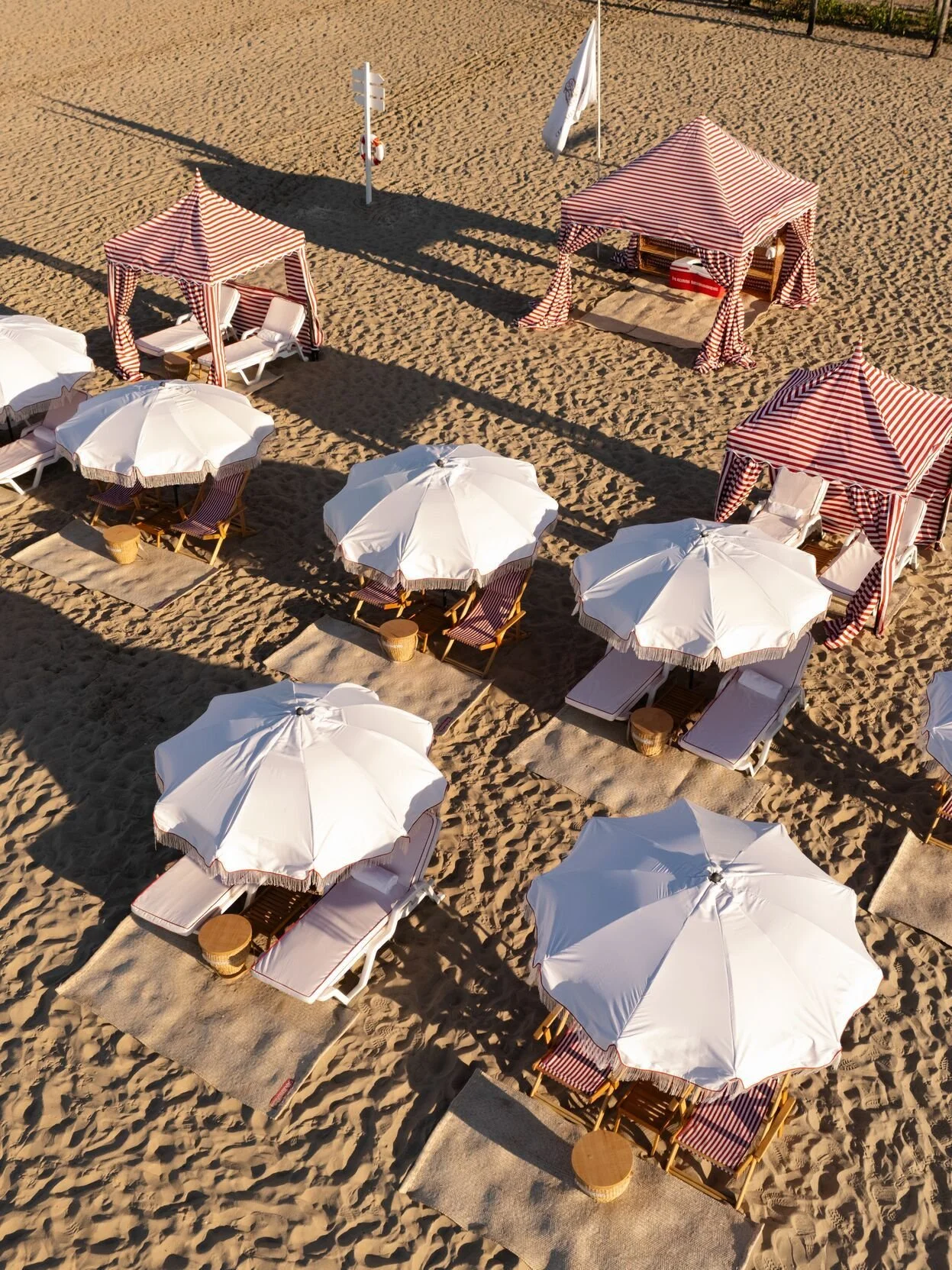 Beach setup with white umbrellas, wooden deck chairs with red and white striped cushions, and two red and white striped cabanas lined up on sandy beach.