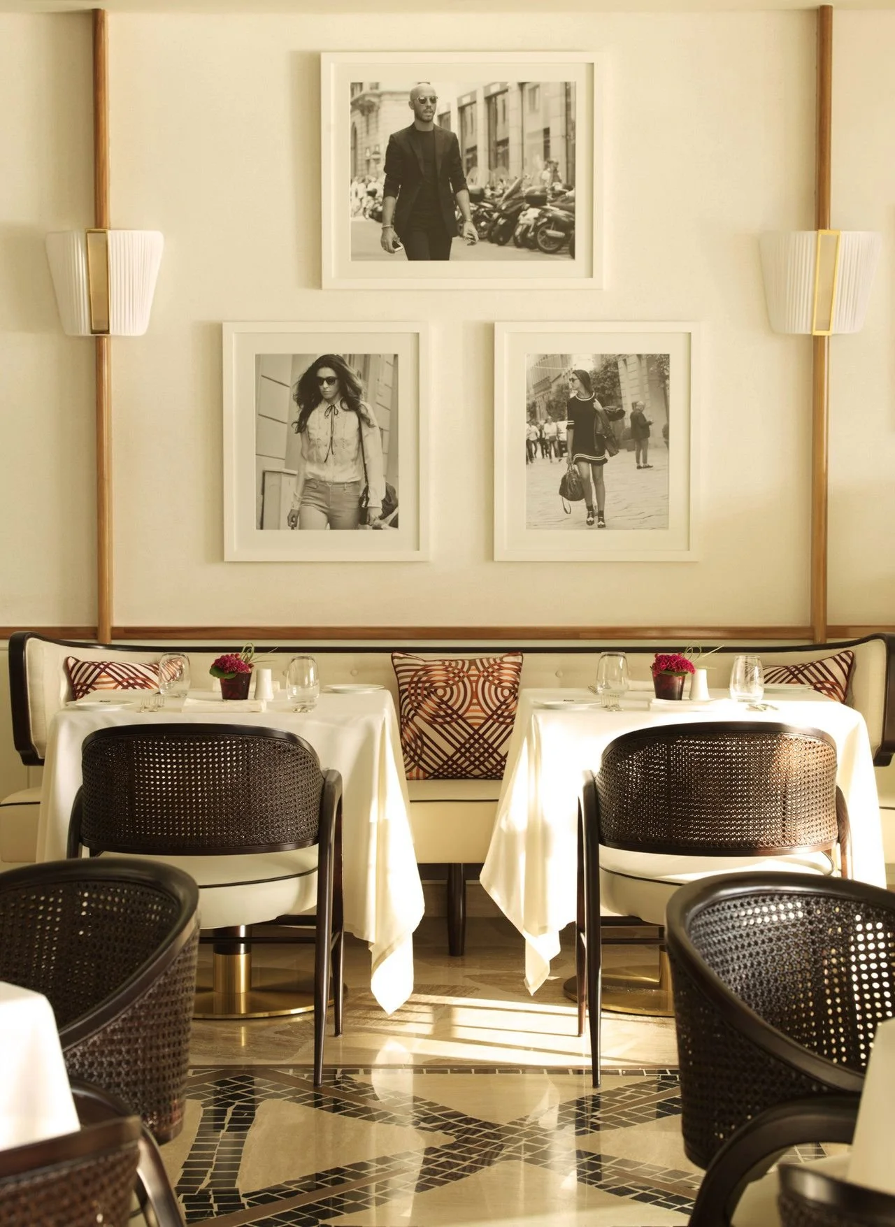 Elegant restaurant dining area with tables set with white tablecloths, glasses, and small potted plants. Dark woven chairs surround the tables. On the wall, framed black-and-white photographs of people walking in city streets are displayed.
