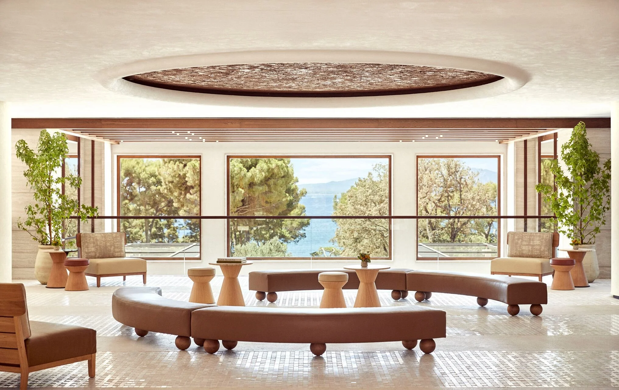 A modern living room with large windows overlooking trees and water. The room has minimalist furniture including a curved beige sofa, matching chairs, and wooden side tables, with potted plants for decoration.