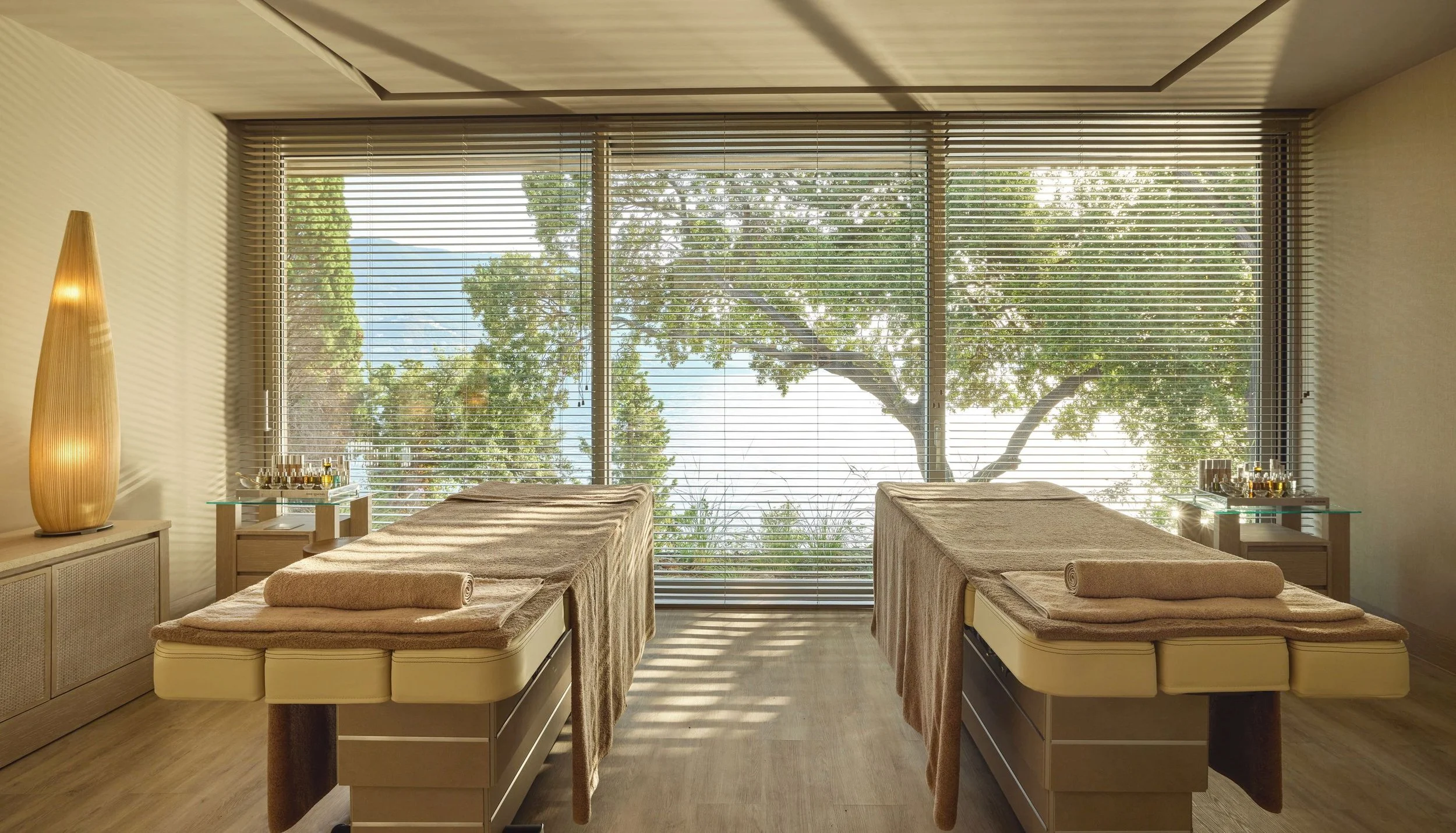 A massage or treatment room with two massage tables covered in beige towels, large windows with blinds showing trees outside, a tall wooden lamp on the left, and small tables with spa products on both sides.