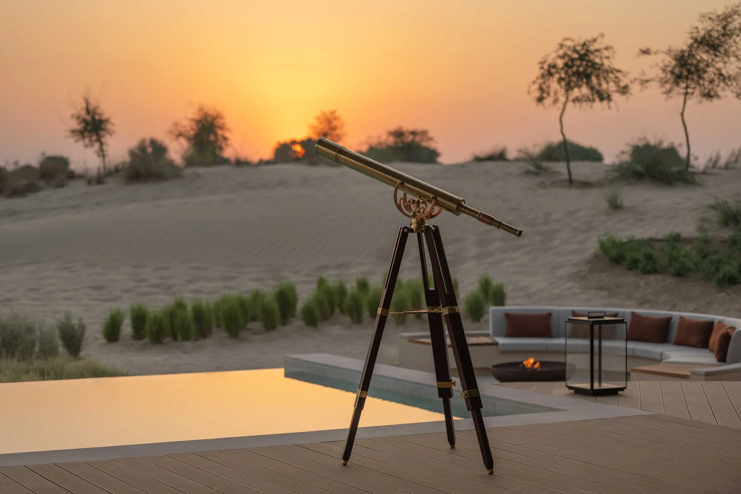 A telescope set up outdoors on a deck during sunset, overlooking a desert landscape with trees and sand dunes.