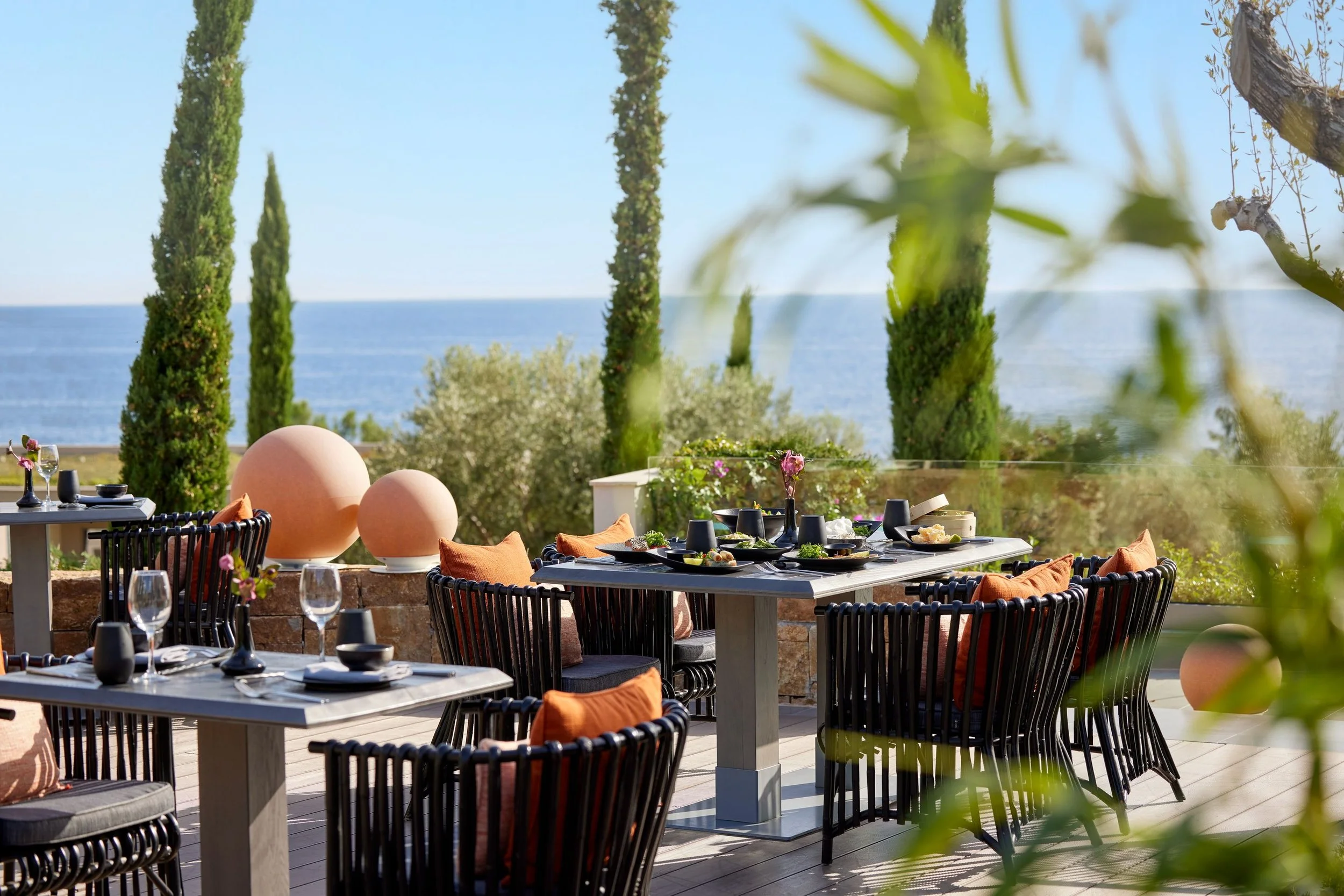 Outdoor dining area with tables set with black dishes, glassware, and orange pillows on black woven chairs, overlooking a body of water with tall green trees and clear sky.