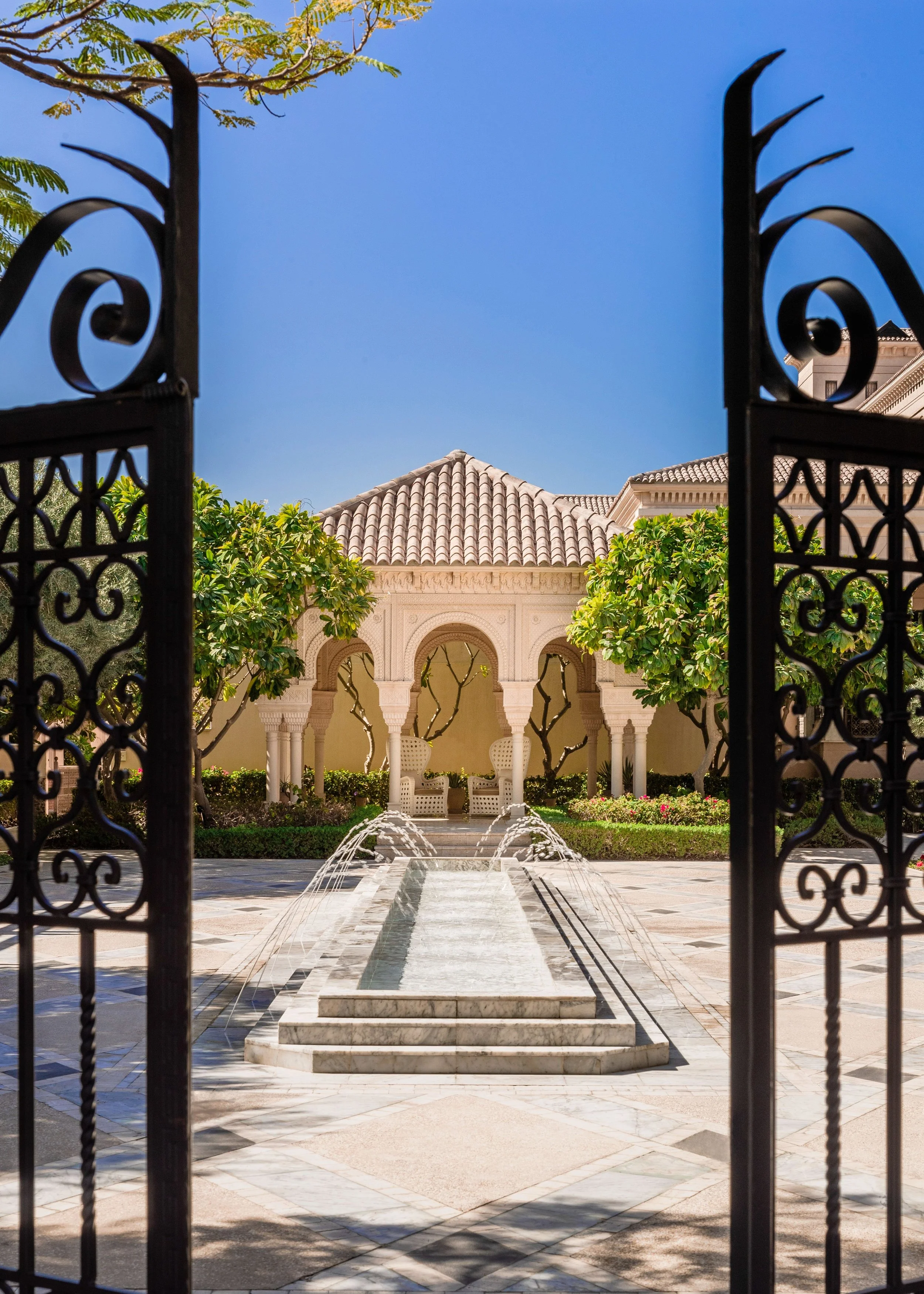 View of a courtyard with a marble fountain, surrounded by green trees and a building with beige walls and tiled roofs, seen through an ornate black wrought iron gate. Blue sky and a few tree branches are visible above.