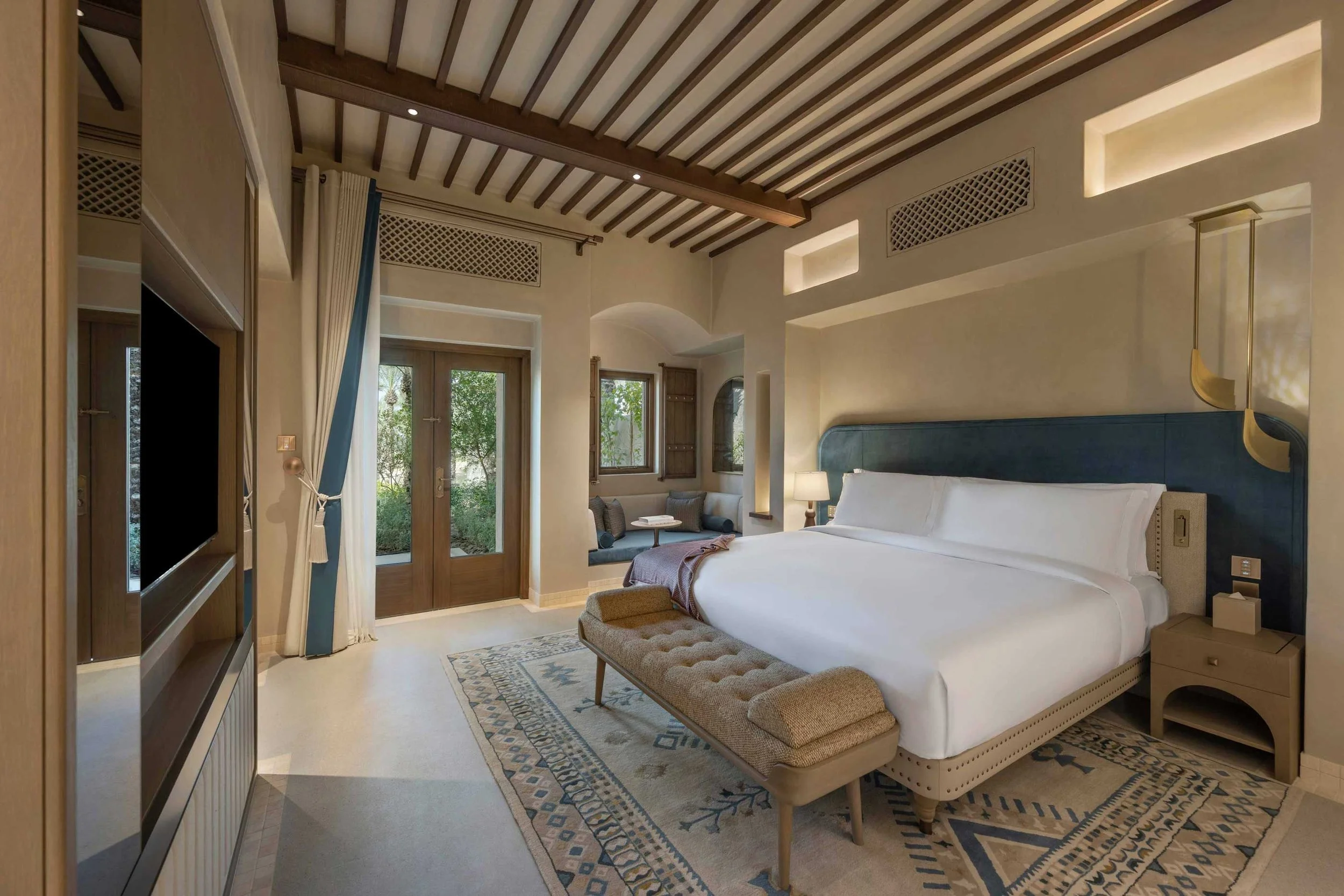 A spacious bedroom with a large bed, a patterned area rug, a bench at the foot of the bed, a side table with a lamp, and a sitting area near a window with wooden shutters, wood-beamed ceiling, and glass doors leading outside.