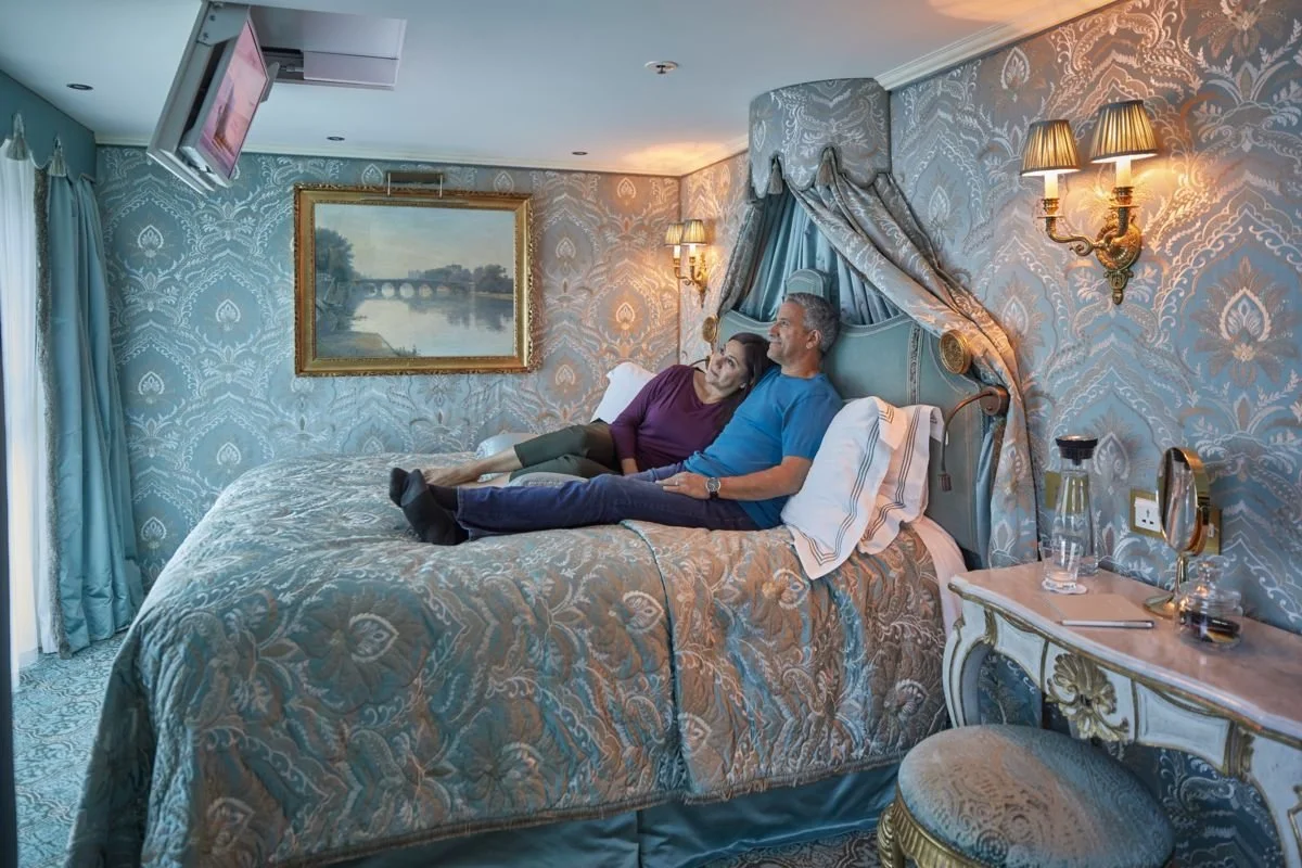 A middle-aged couple lounged on a bed in a lavish decorated bedroom, watching TV. The room has elegant wallpaper, a painting of a river scene, and ornate lamp fixtures.